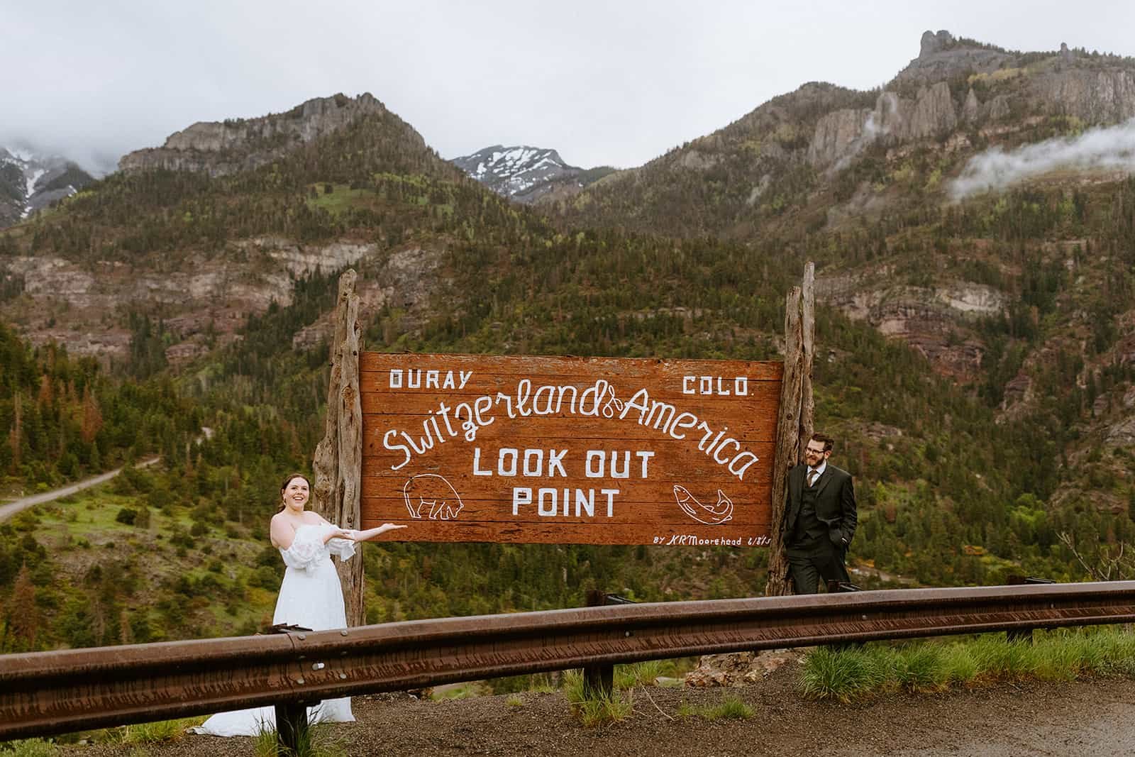 A couple in wedding clothes stands in front of the Switzerland of America sign in Ouray, Colorado