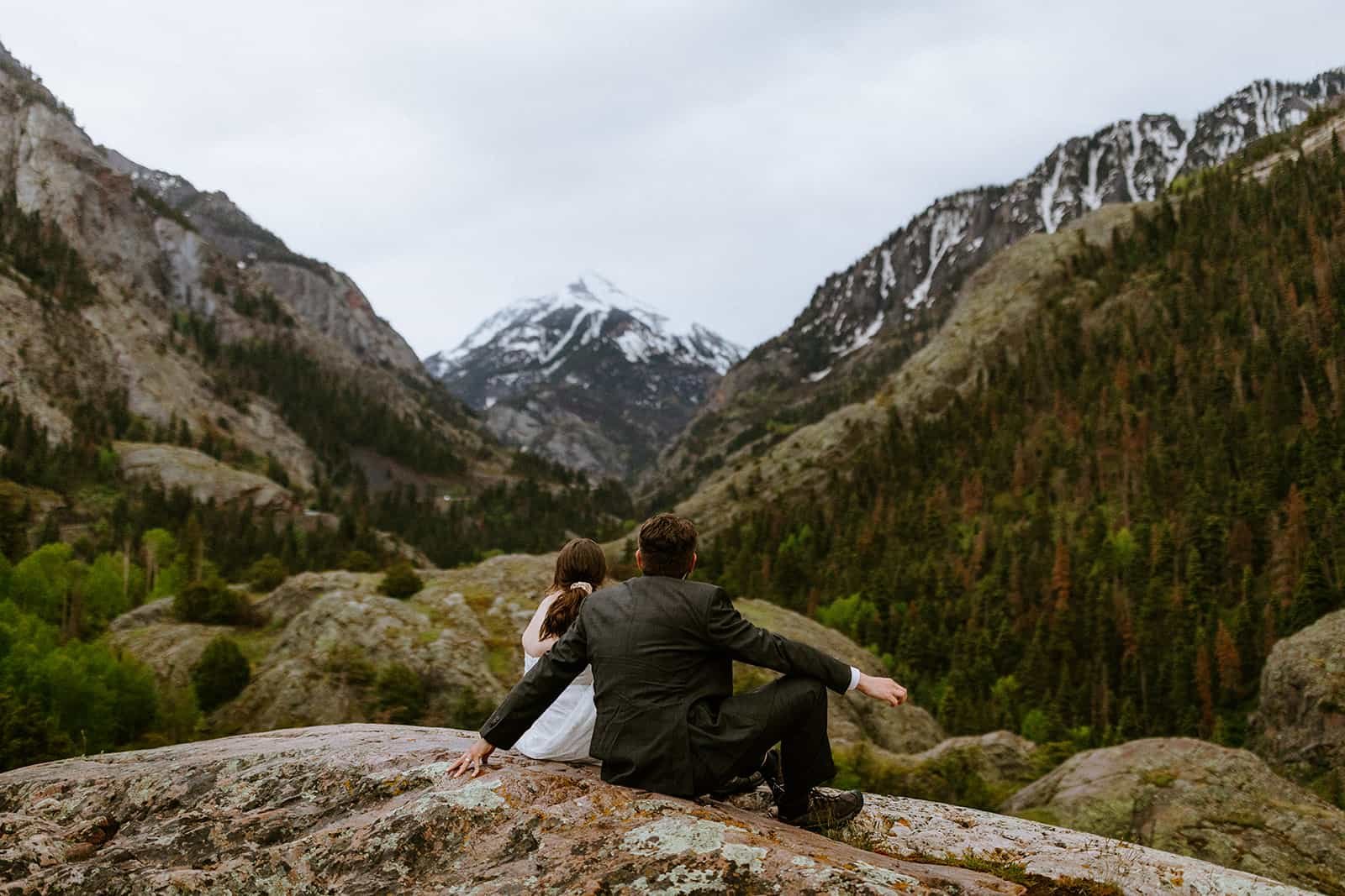 A couple sits down on a wet rock in their wedding clothes looking at the mountains