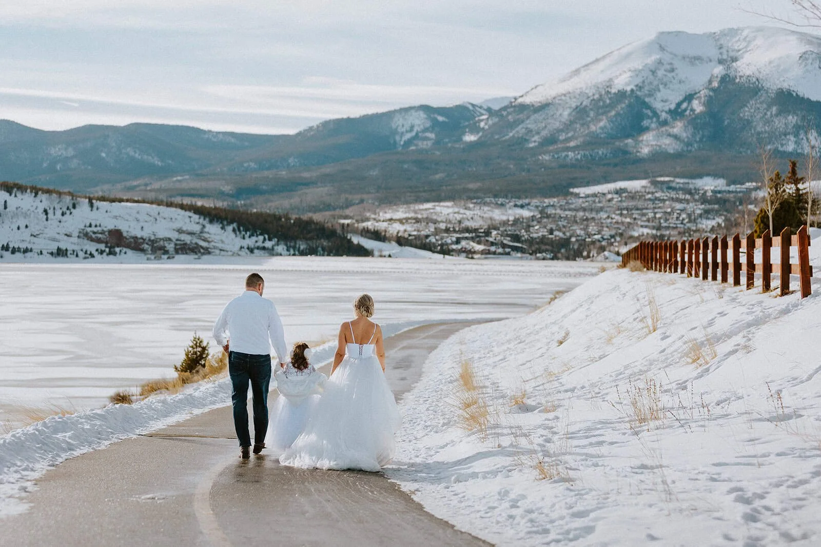 A couple in wedding clothes walks down a snowy road above a frozen Lake Dillon