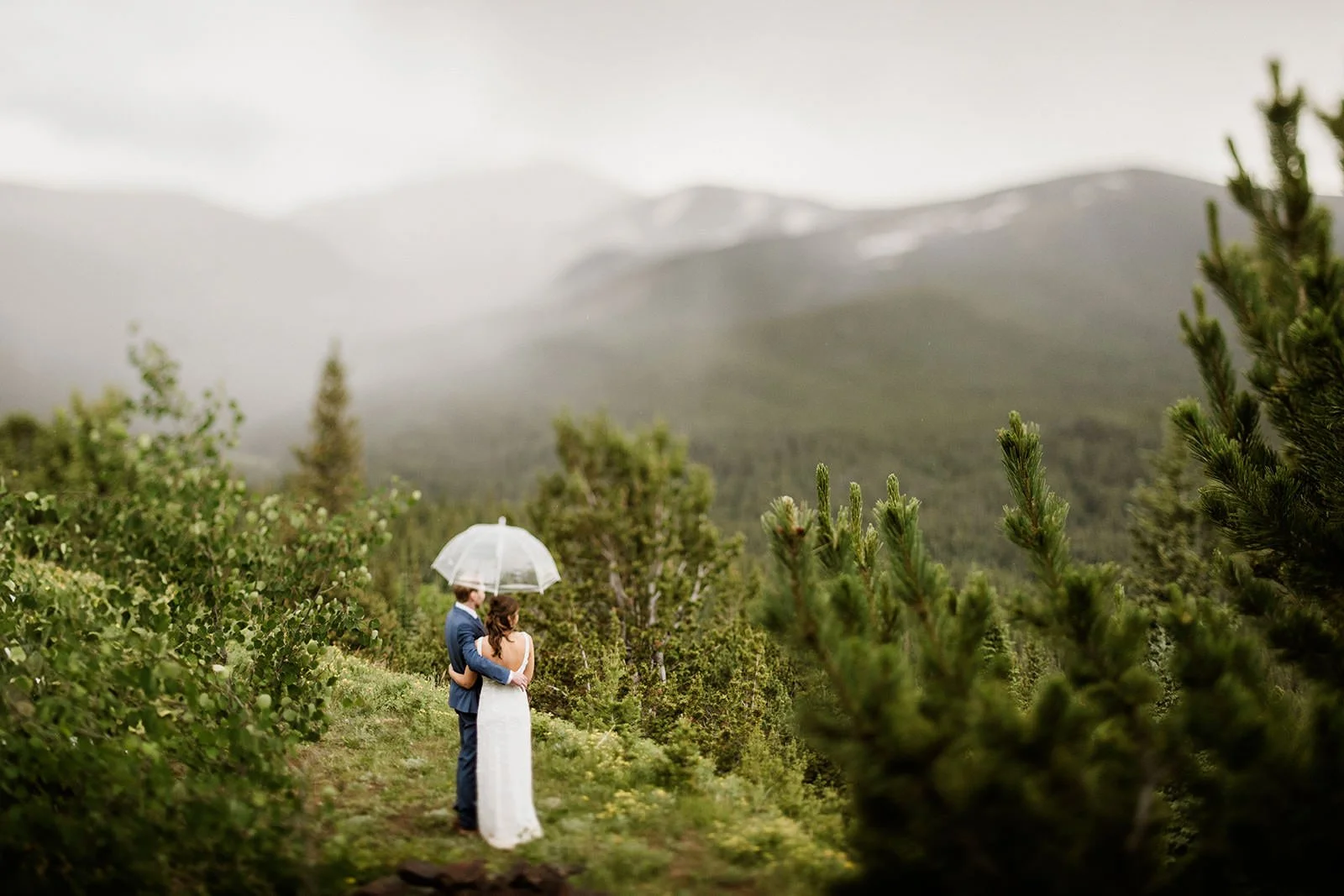 A couple stands under a clear umbrella looking over a foggy Colorado landscape