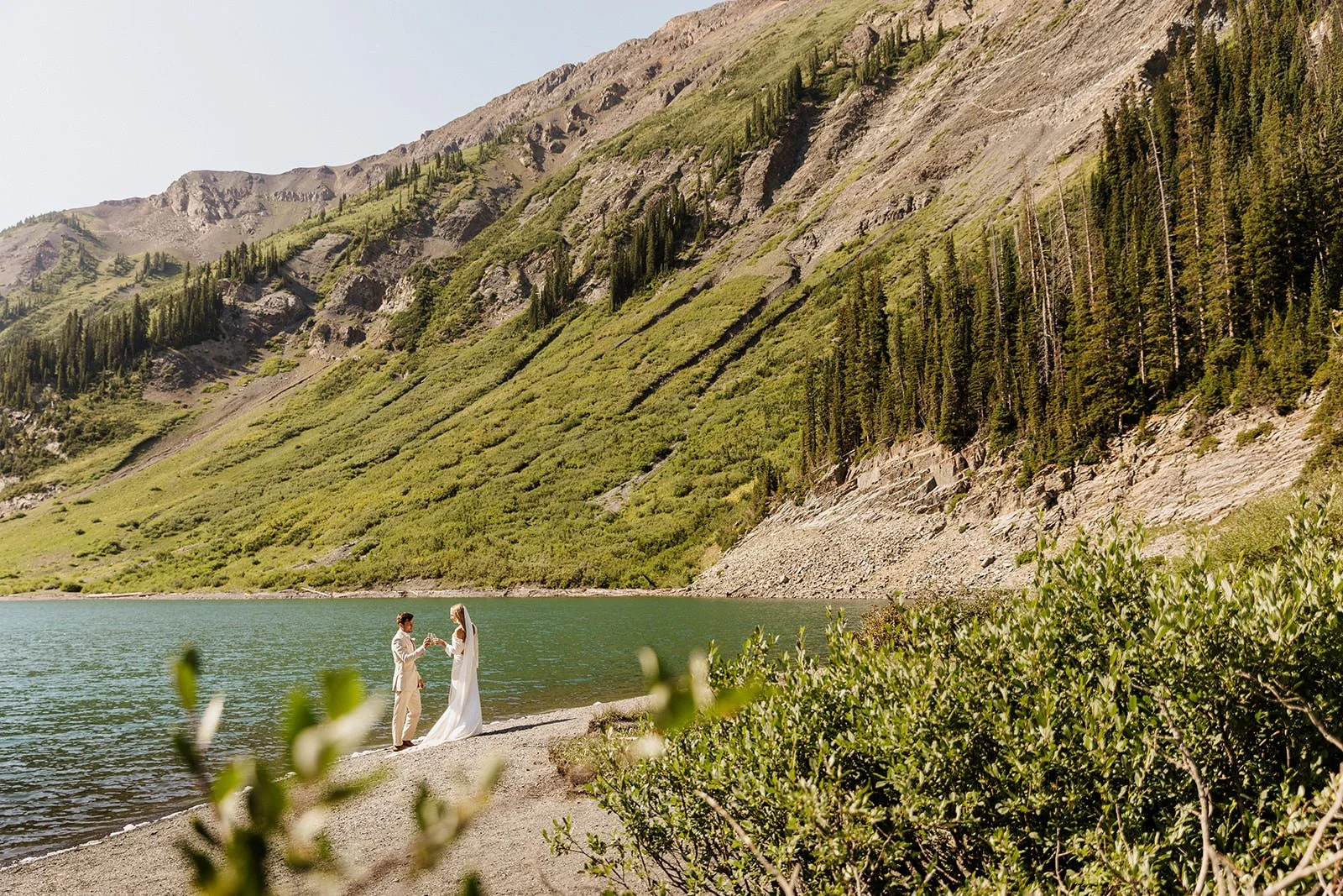 Bride and groom in front of a blue alpine lake and tall green mountains