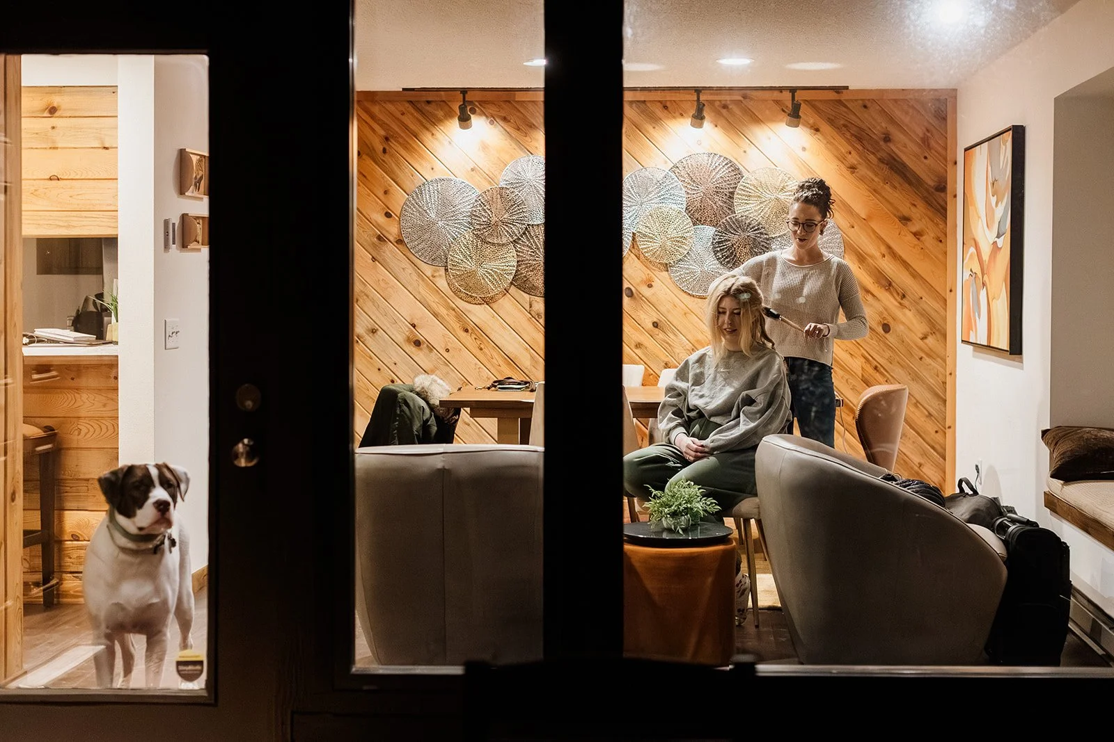 Bride having her hair done inside a cabin in Estes Park at night while her dog waits at the glass door