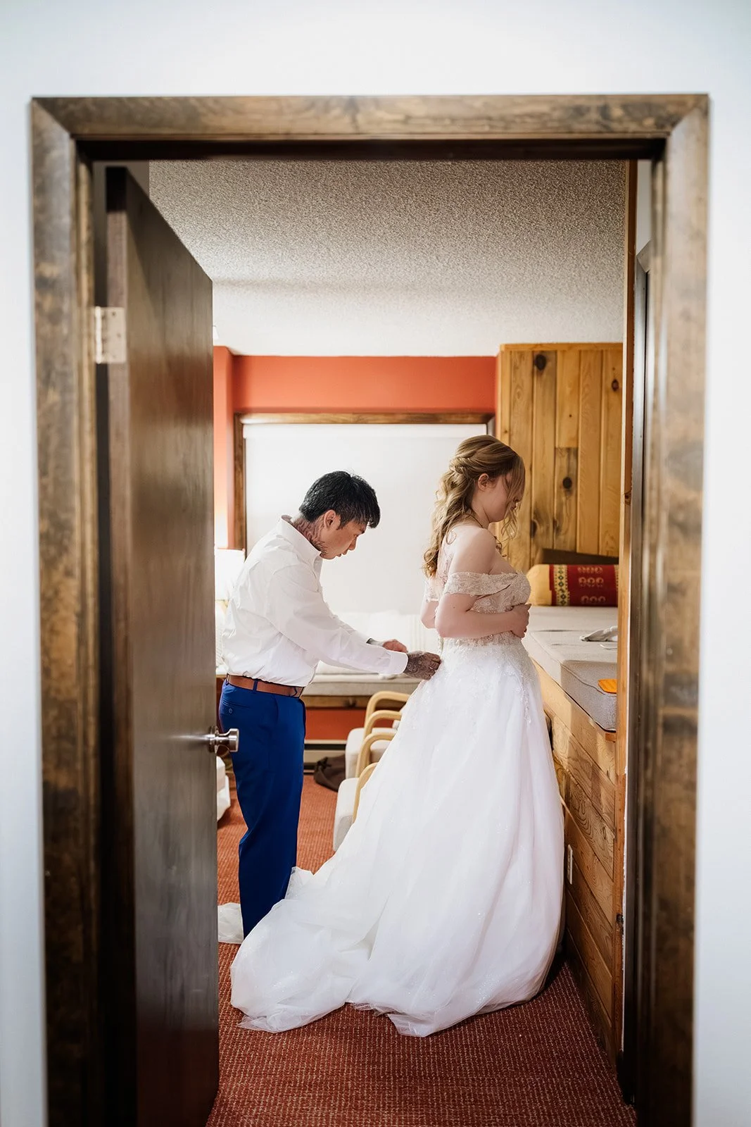 Groom buttoning the back of the bride's wedding dress, seen through a bedroom doorway