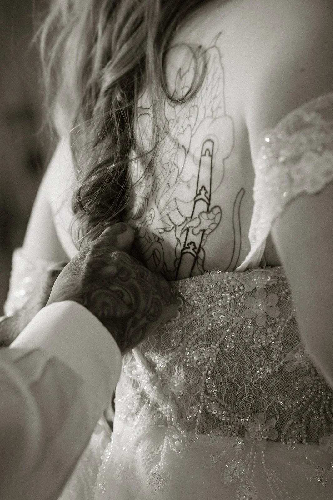 Close up of hands fastening a lace wedding dress over the bride's back tattoo, in black and white