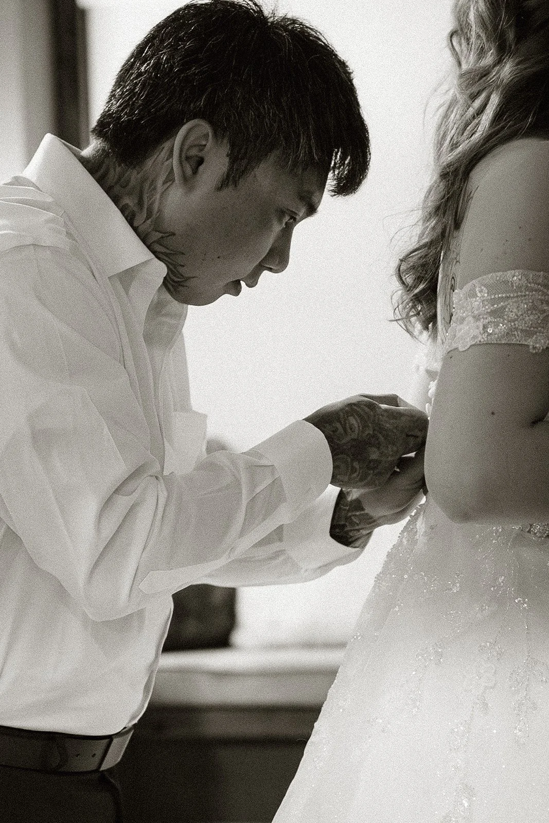 Groom in a white shirt concentrating on the buttons of the bride's dress, in black and white