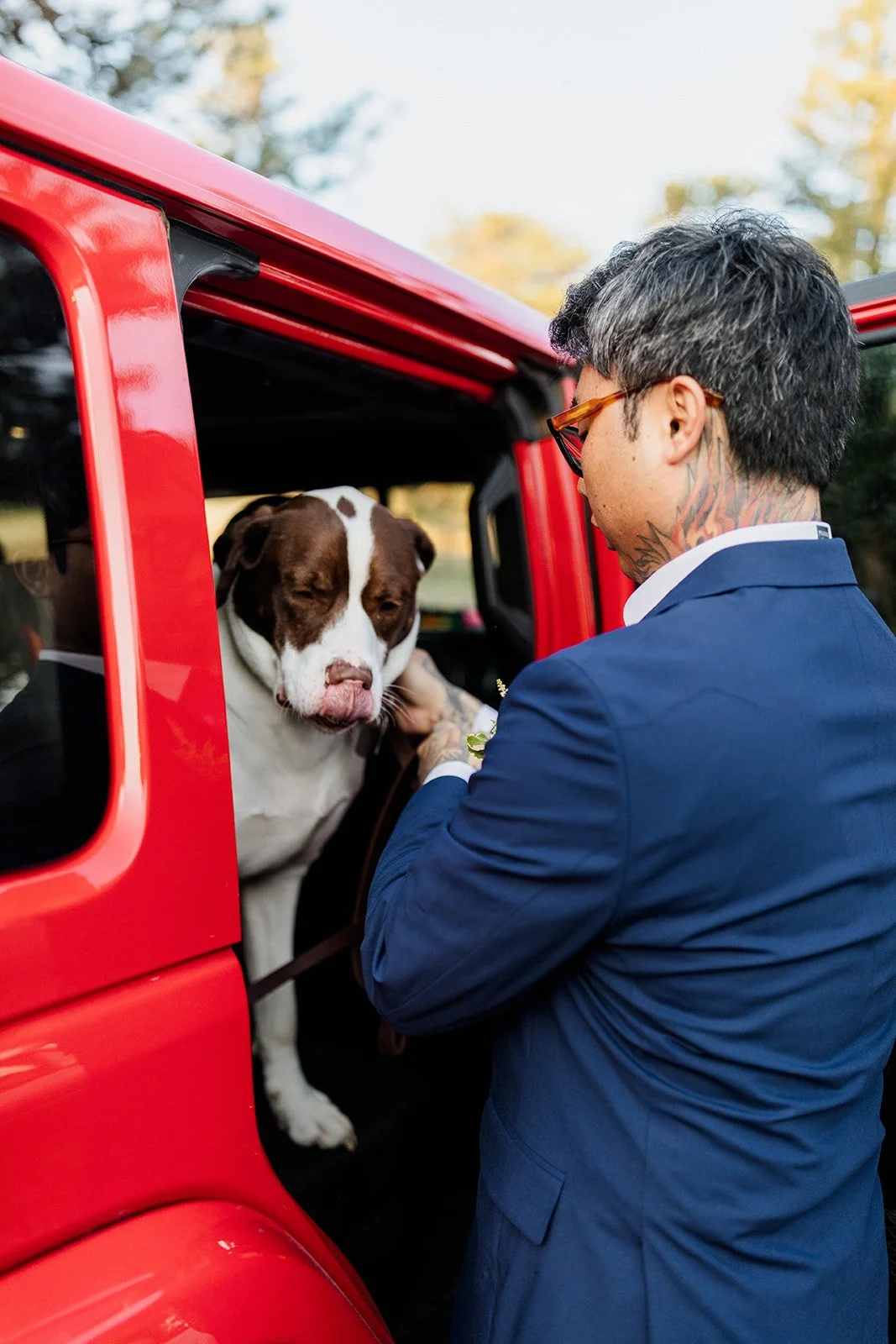 Groom greeting his dog as it leans out of the door of a red Jeep