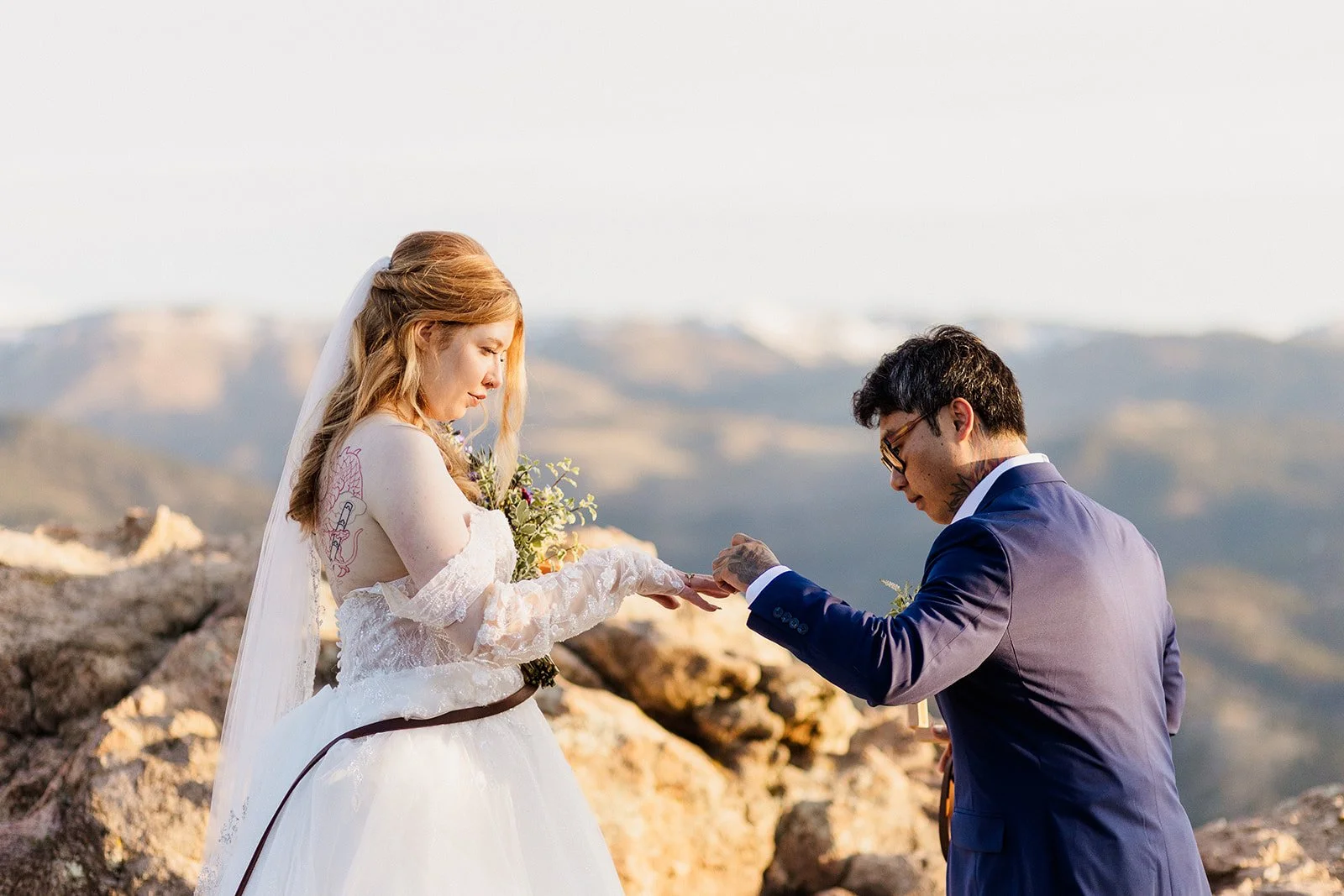 Groom holding the bride's hand during their ring exchange with the foothills behind them