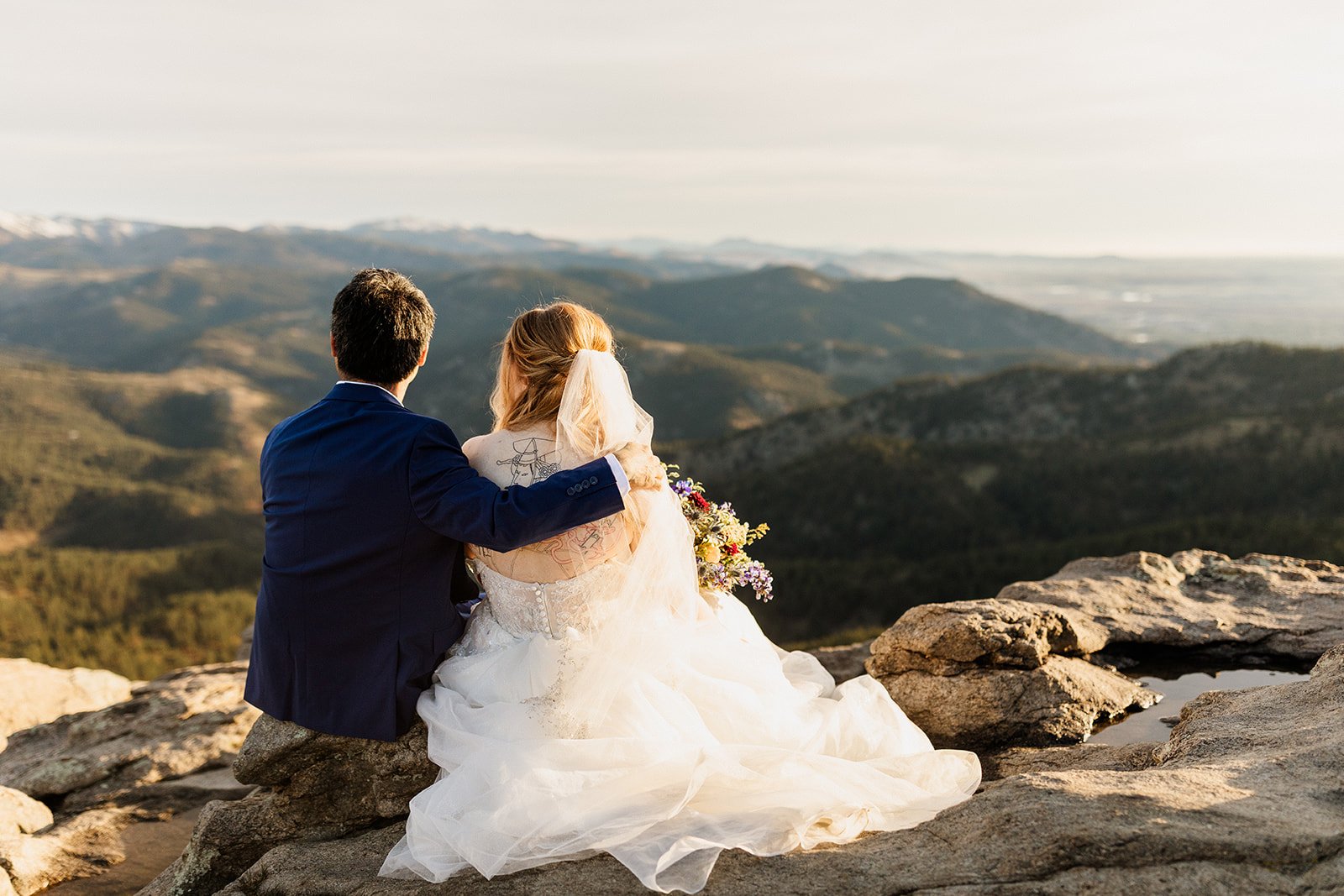 Couple sitting on a rock ledge with his arm around her, looking out over the foothills
