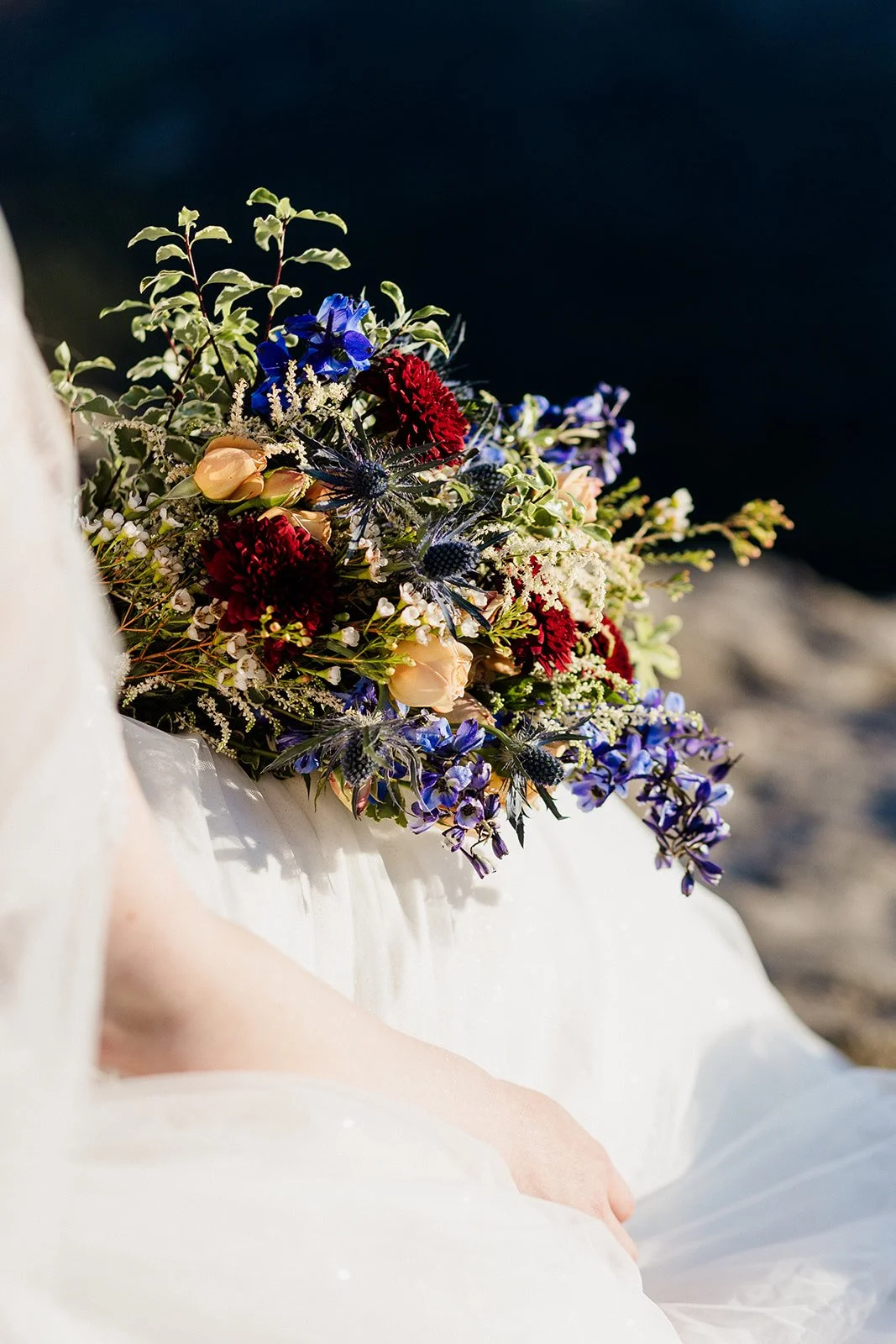 Wildflower bouquet with deep red, blue and peach flowers resting on the bride's lap