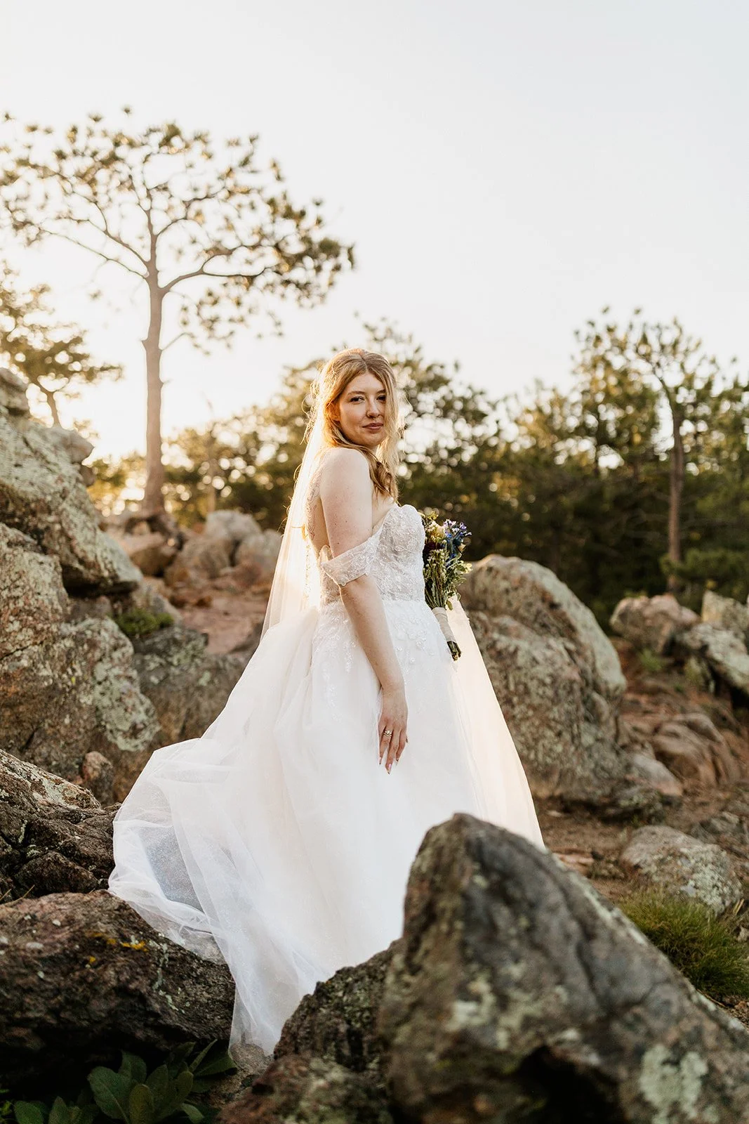 Bride in a lace wedding dress and veil standing on the rocks in golden light