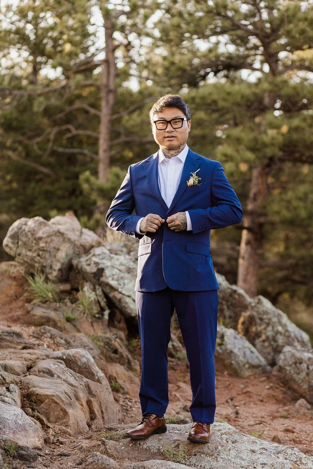 Groom buttoning his blue suit jacket on the rocks under the pines