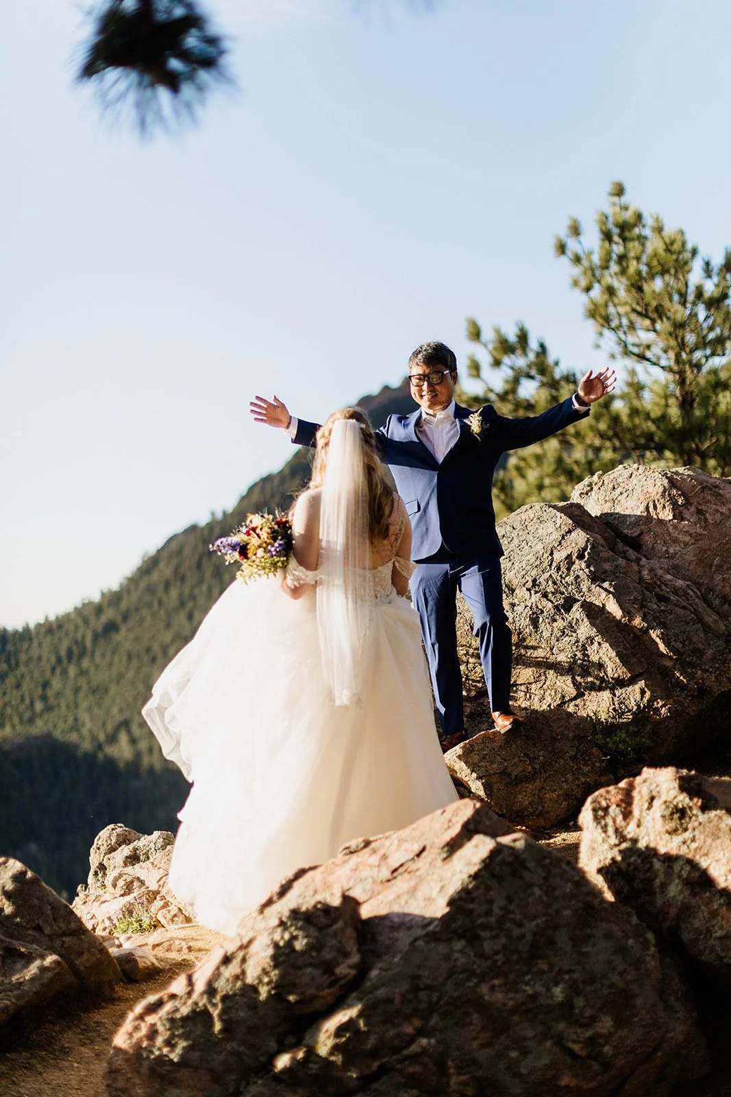 Groom throwing his arms out on top of a boulder while the bride stands below