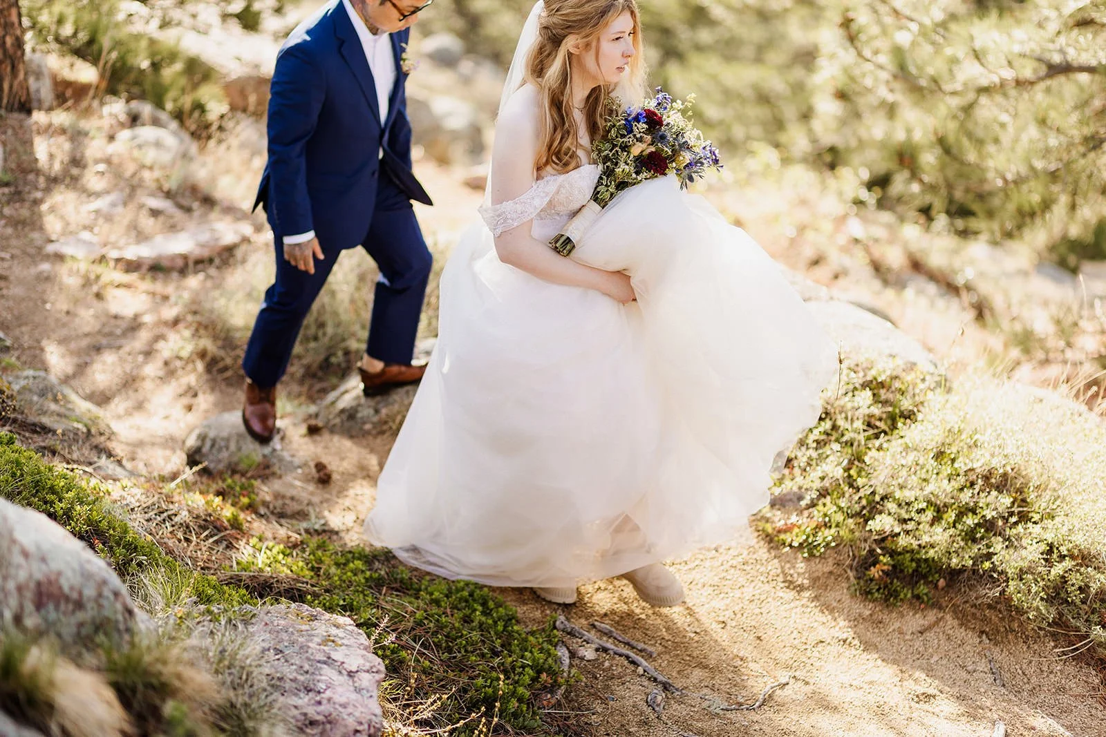 Bride carrying her dress down a rocky trail with the groom behind her