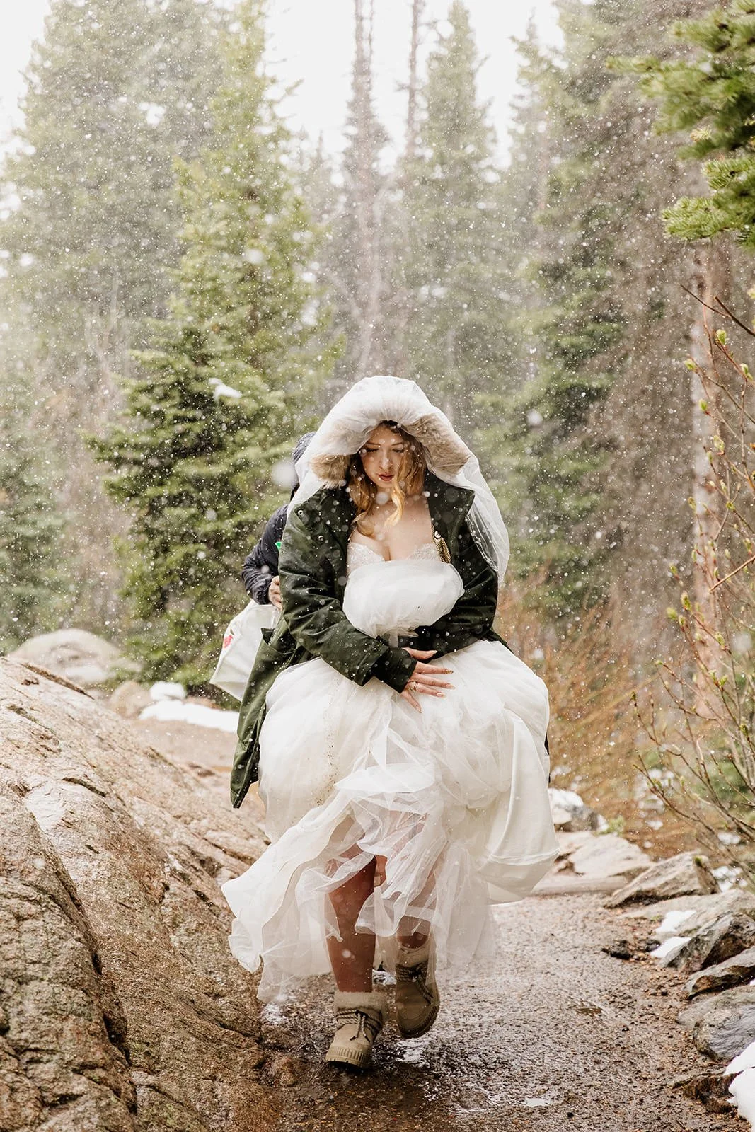 Bride hiking to Dream Lake in falling snow with a coat over her wedding dress