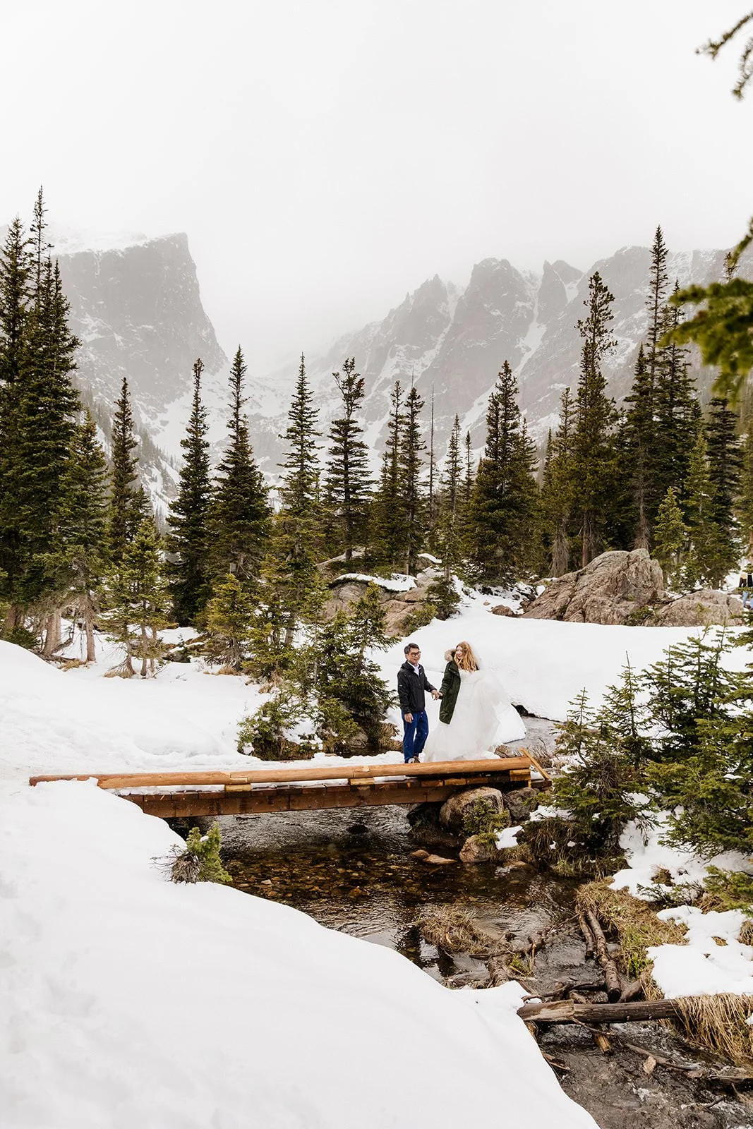 Couple holding hands on a snowy footbridge with Hallett Peak behind them