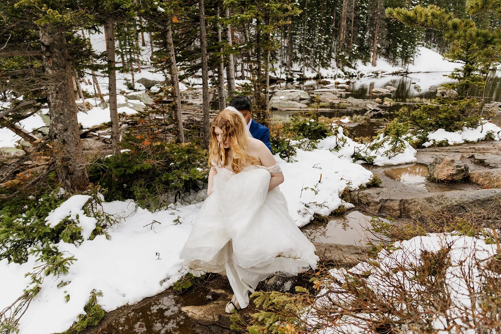 Bride lifting her dress to step across a stream in the snow