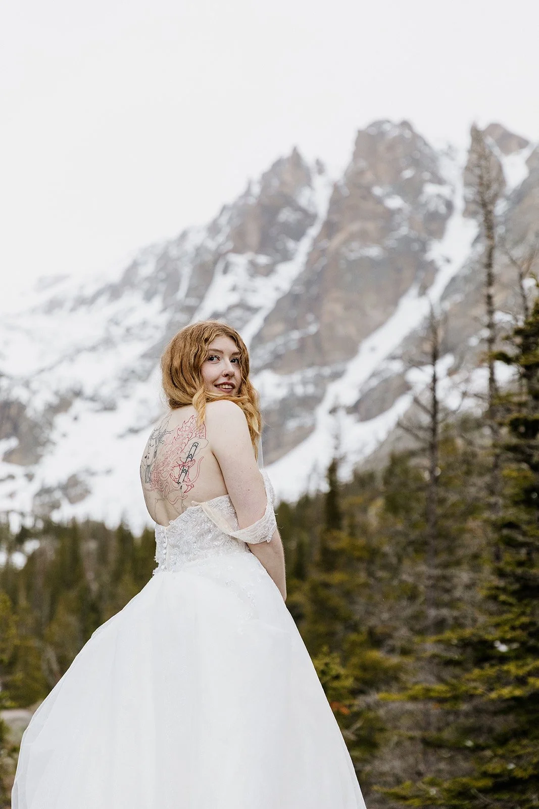 Bride looking back over her shoulder with snow covered peaks behind her and her back tattoo piece showing