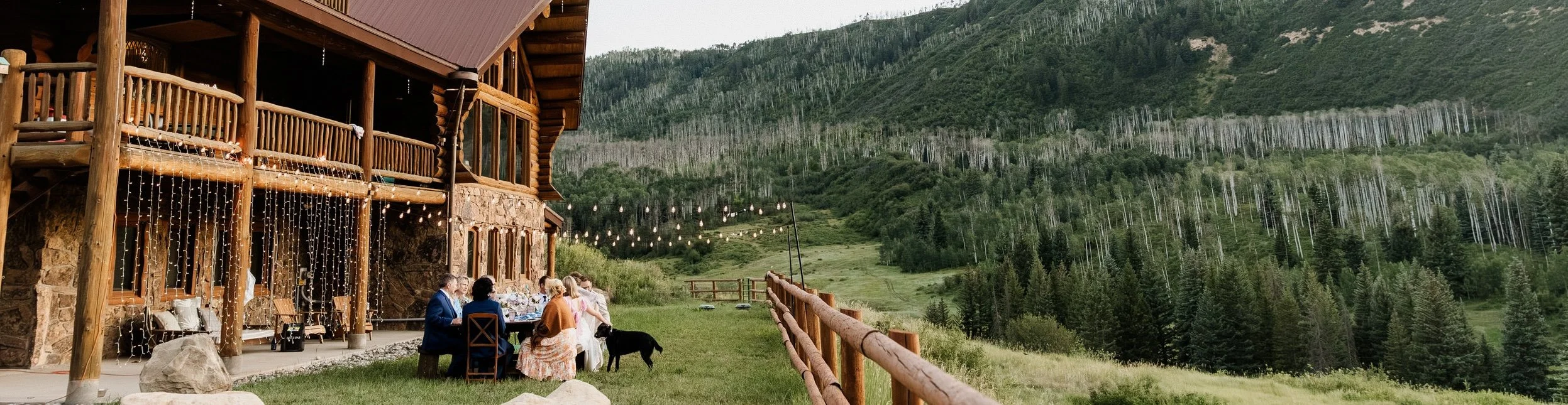 People sitting at a table outside a rustic lodge with string lights, overlooking mountains covered in trees and grassy slopes.