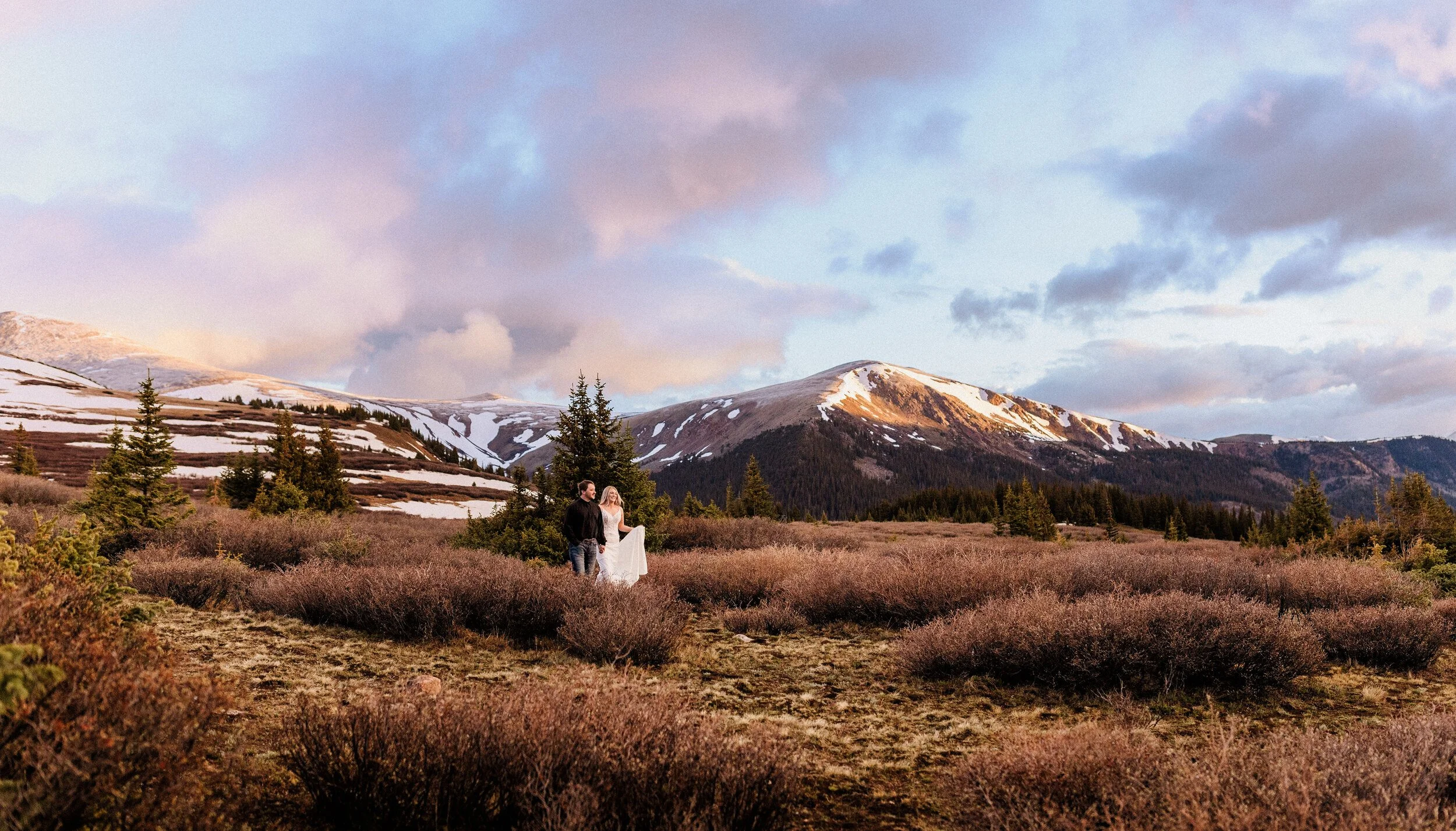 A bide and groom walk hand in hand in front of a pink Colorado sunrise outside of Georgetown.