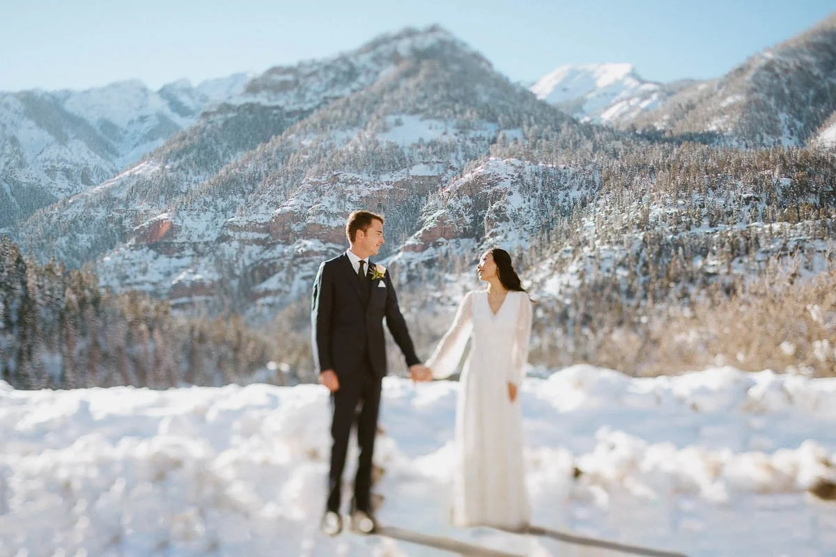 A couple stands side by side holding hands in wedding attire outside of Ouray in the winter with snow covered mountains behind them