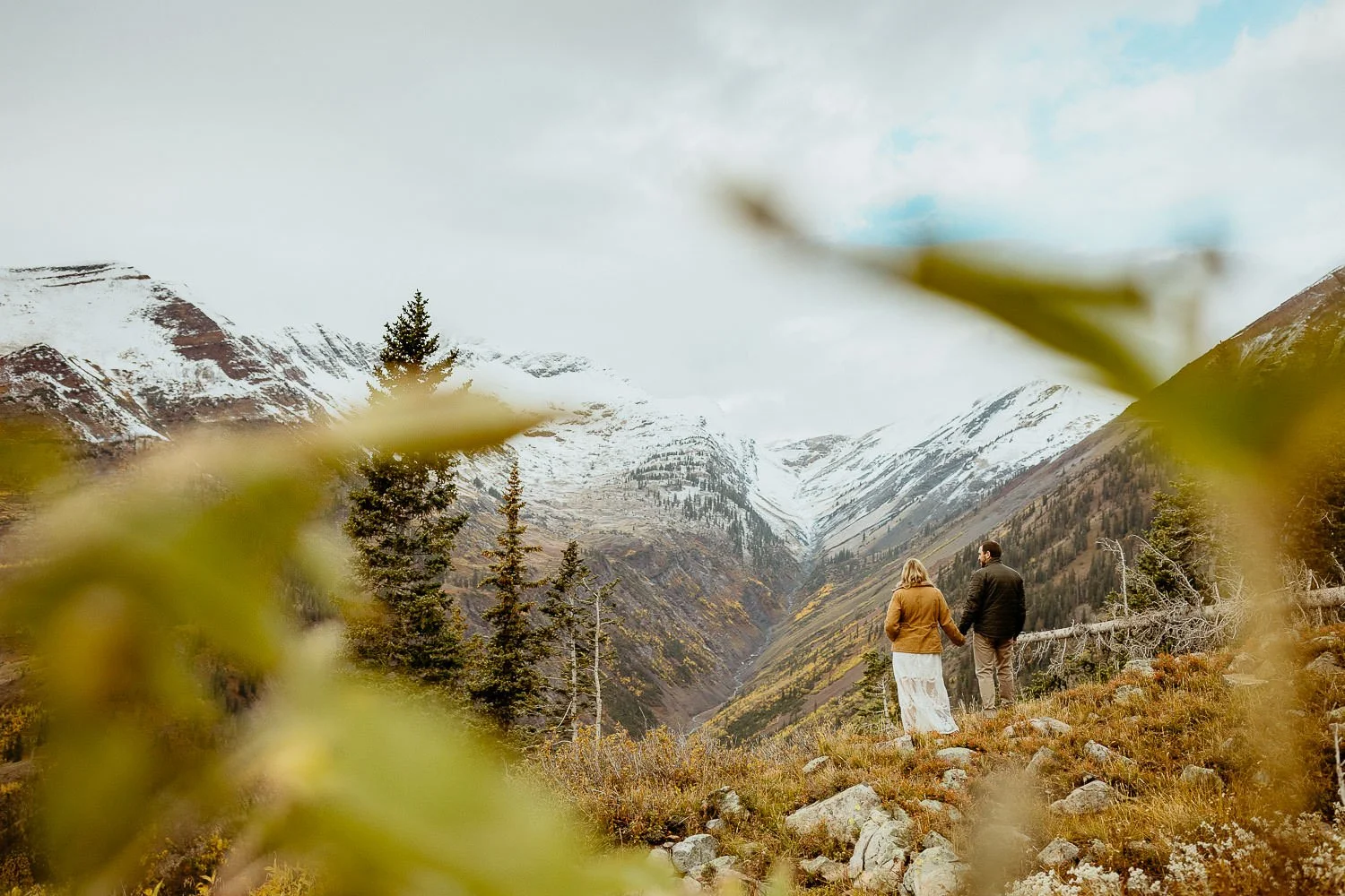 A couple stands on a hillside in Crested Butte with snow on the peaks behind them