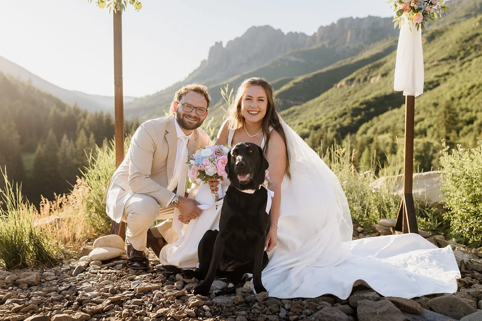 A newlywed couple with a service dog posing outdoors with mountains in the background. The bride is in a white wedding dress holding a bouquet, and the groom is in a light-colored suit. The black Labrador retriever is sitting in front of them. The scene is bright with sunlight.