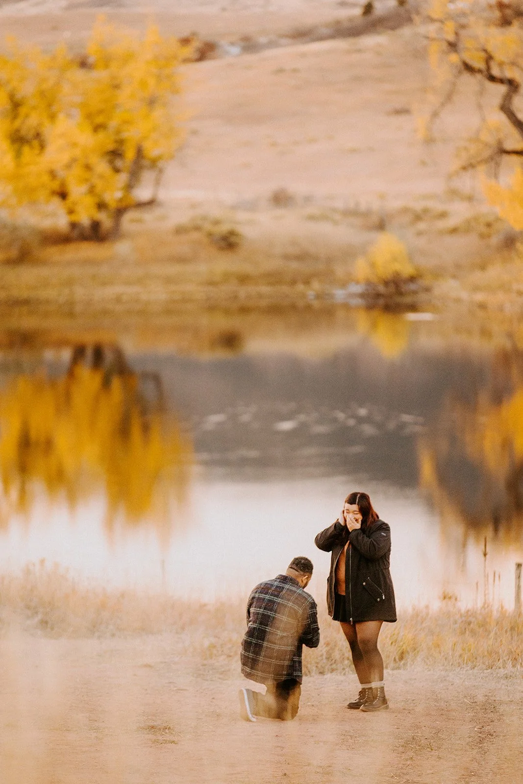A surprised woman covering her face as her partner kneels by a pond lined with golden fall trees