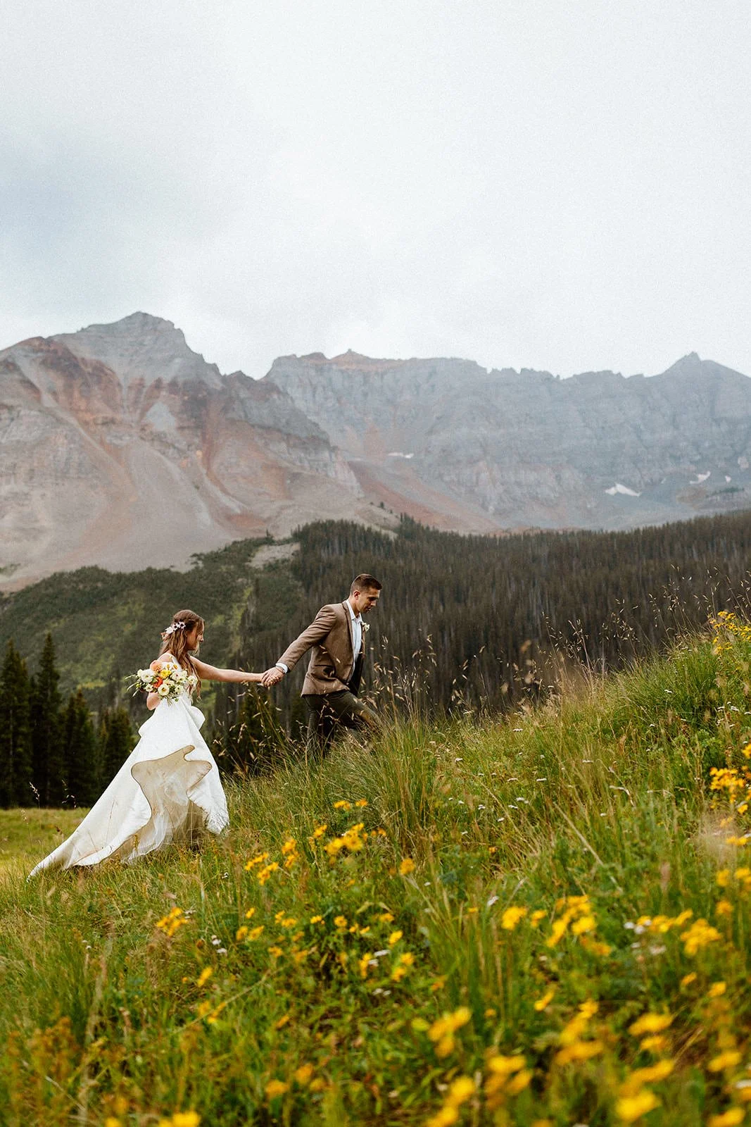 A beautiful Telluride elopement scene featuring a couple holding hands as they run through wildflowers and lush greenery, with majestic mountains in the background. This romantic jeep elopement captures a unique Colorado adventure.