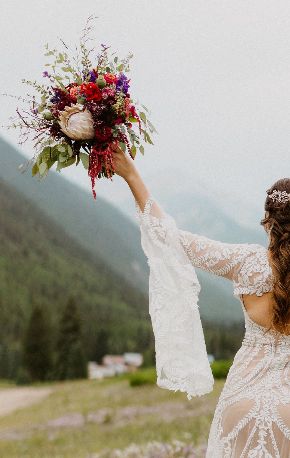 Long sleeve lace elopement dress with bell sleeves, holding a colorful bouquet up toward the mountains