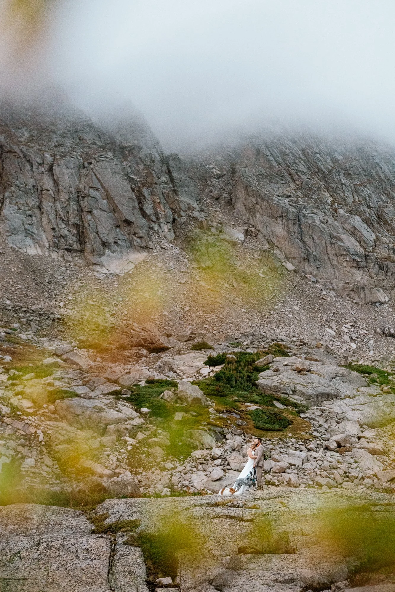 Couple and their two dogs standing small against a huge fog-covered mountain cliff at an alpine lake in Colorado
