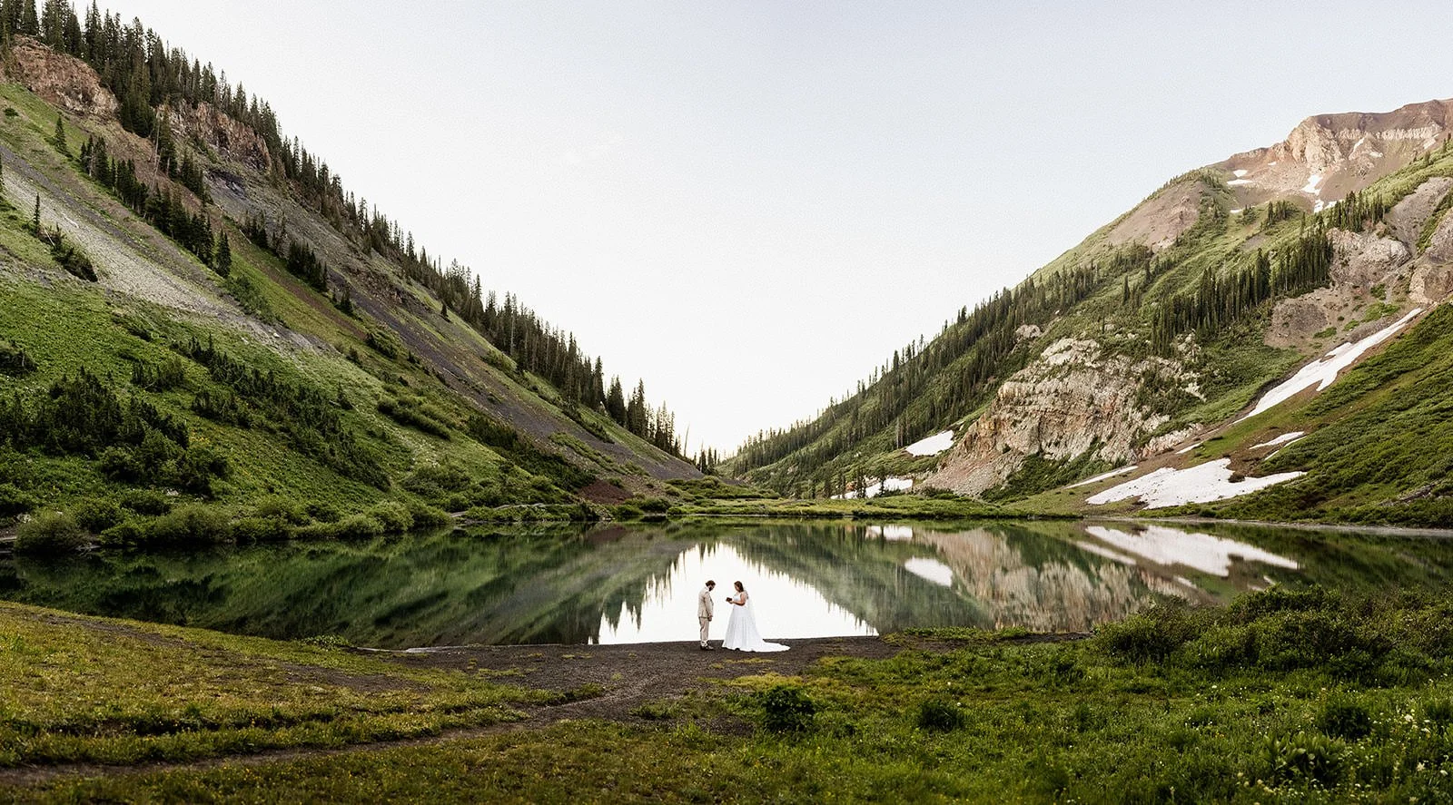 A bride and groom standing together near a calm mountain lake, surrounded by green hills and rocky slopes, with a reflection of the landscape in the water.