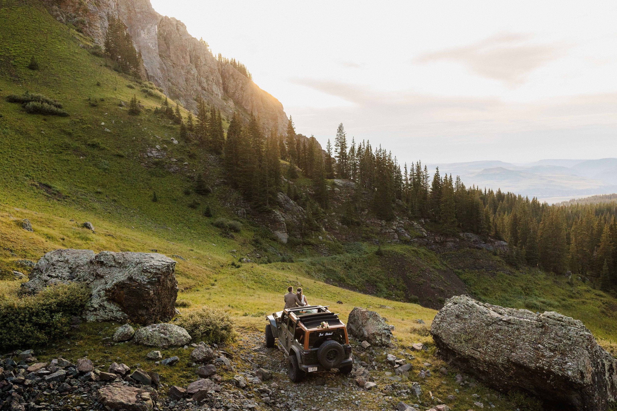 A bride and groom sit on the hood of their jeep watching sunset