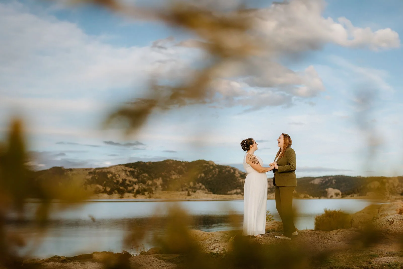Two people holding hands beside a mountain lake, one in a white elopement dress and one in a green suit