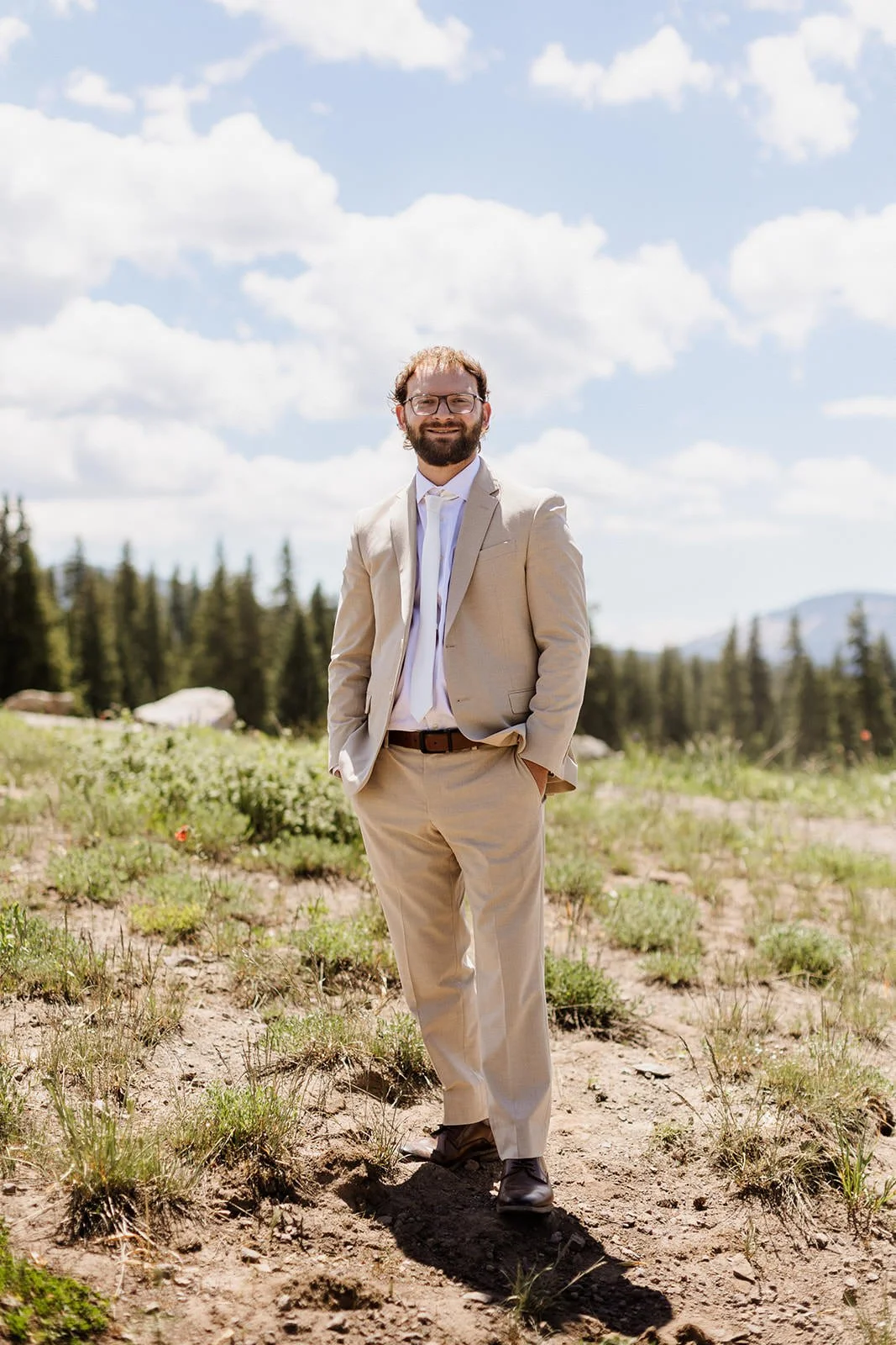 A man wearing a beige suit and glasses standing outdoors in a field with trees and mountains in the background, under a partly cloudy sky.