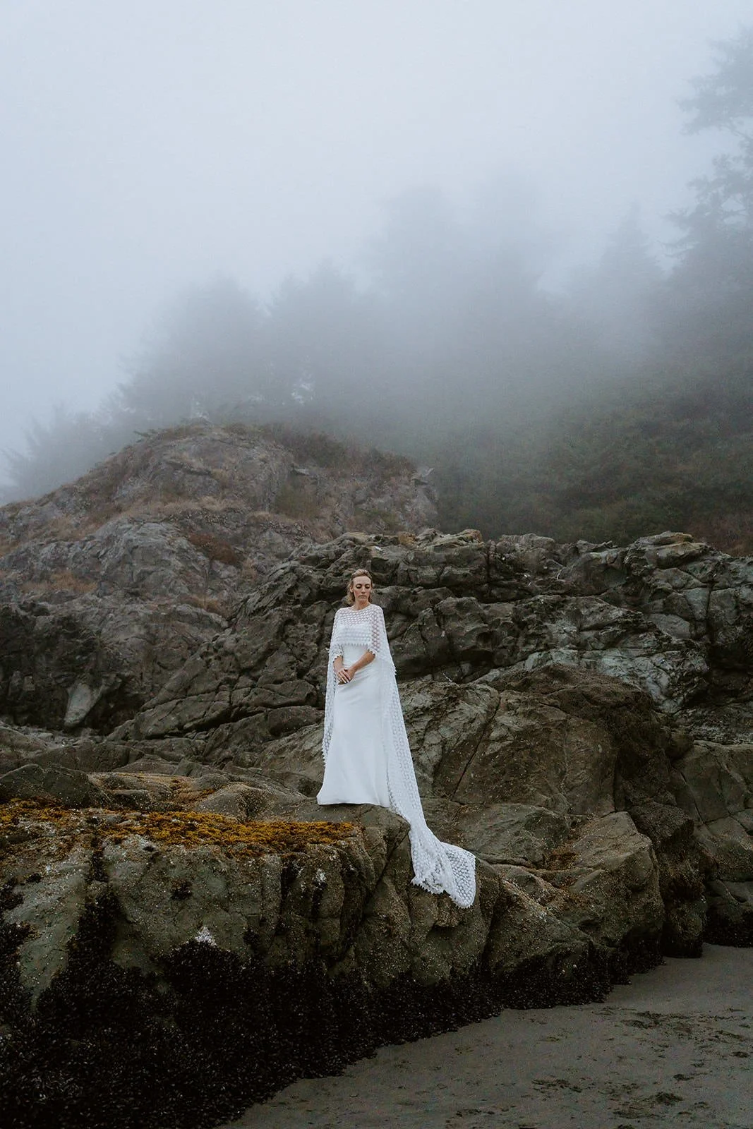 White elopement dress with a long cape, standing on rocks at a foggy beach