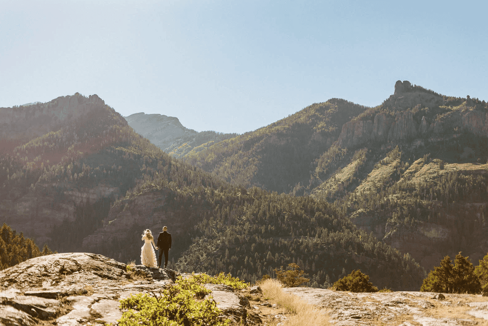 Couple holding hands on a rocky ledge overlooking lush mountain valleys in Ouray, Colorado