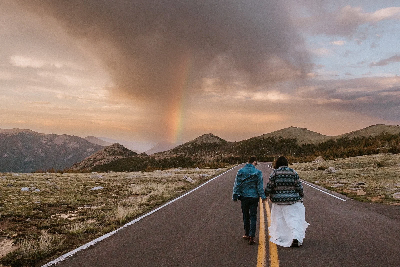 Two people walking hand-in-hand along an empty mountain road at sunset, with a rainbow and cloudy sky above.