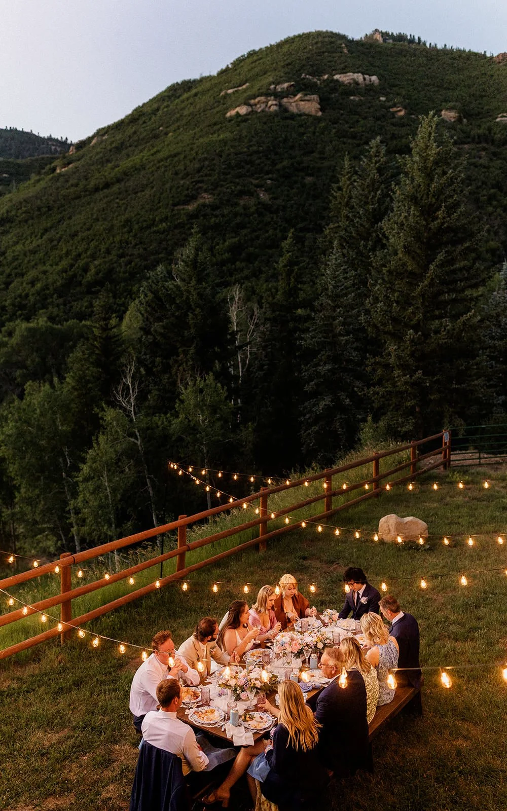 A group of people dining outdoors at a long table decorated with flowers and string lights, set against a backdrop of forested mountains during evening.