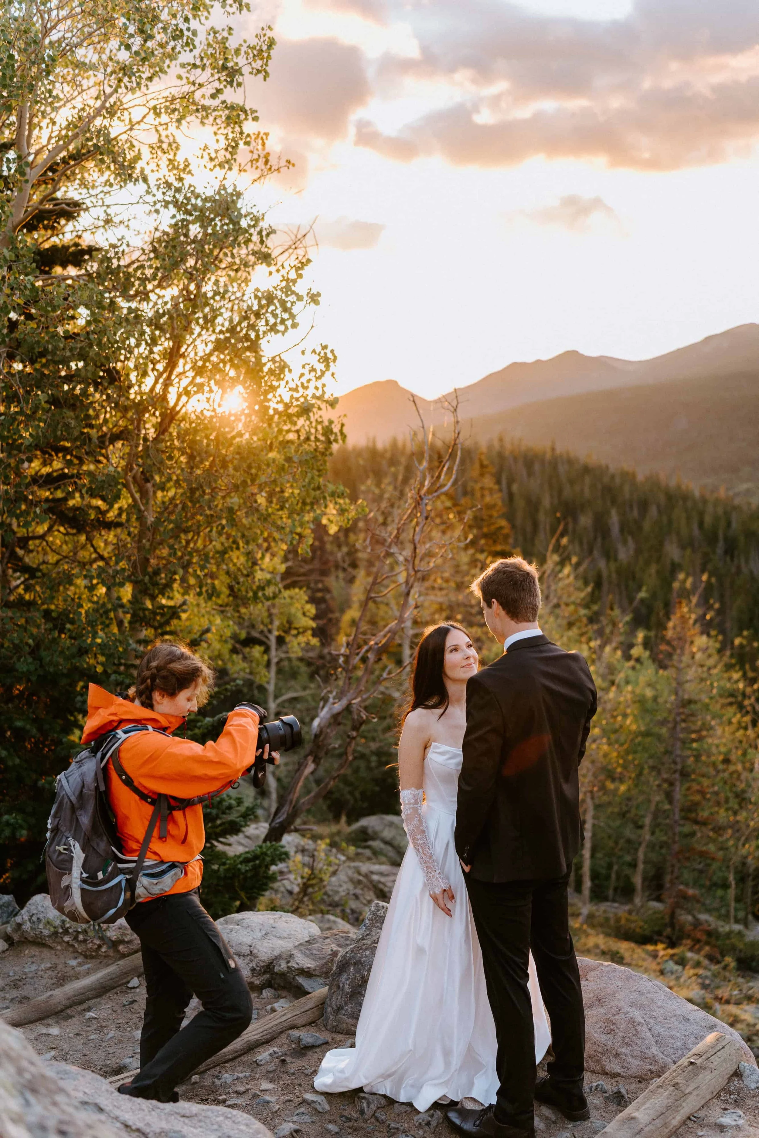 A photographer takes pictures of a bride and groom during a wedding ceremony outdoors at sunset, surrounded by trees and mountains.