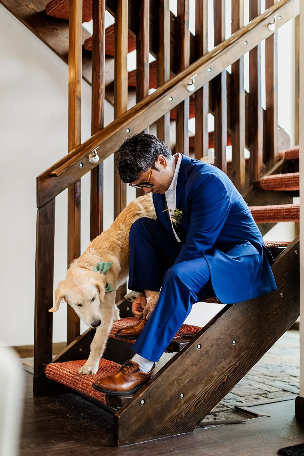 Groom in a blue suit tying his shoes on the cabin stairs before an Estes Park elopement while his dog walks by