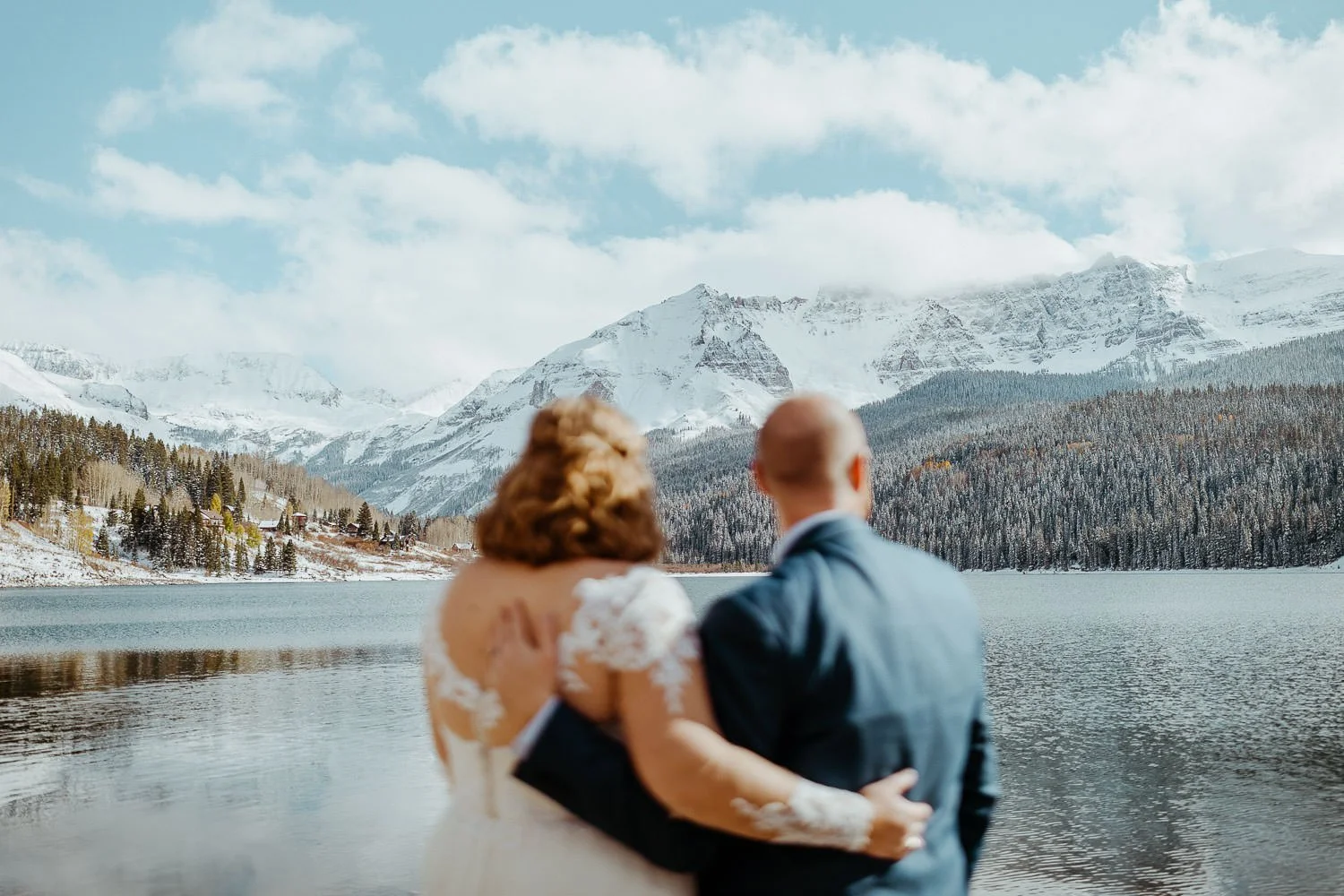 A couple hugs with their backs to the camera, looking across Trout Lake at snowy peaks near Telluride