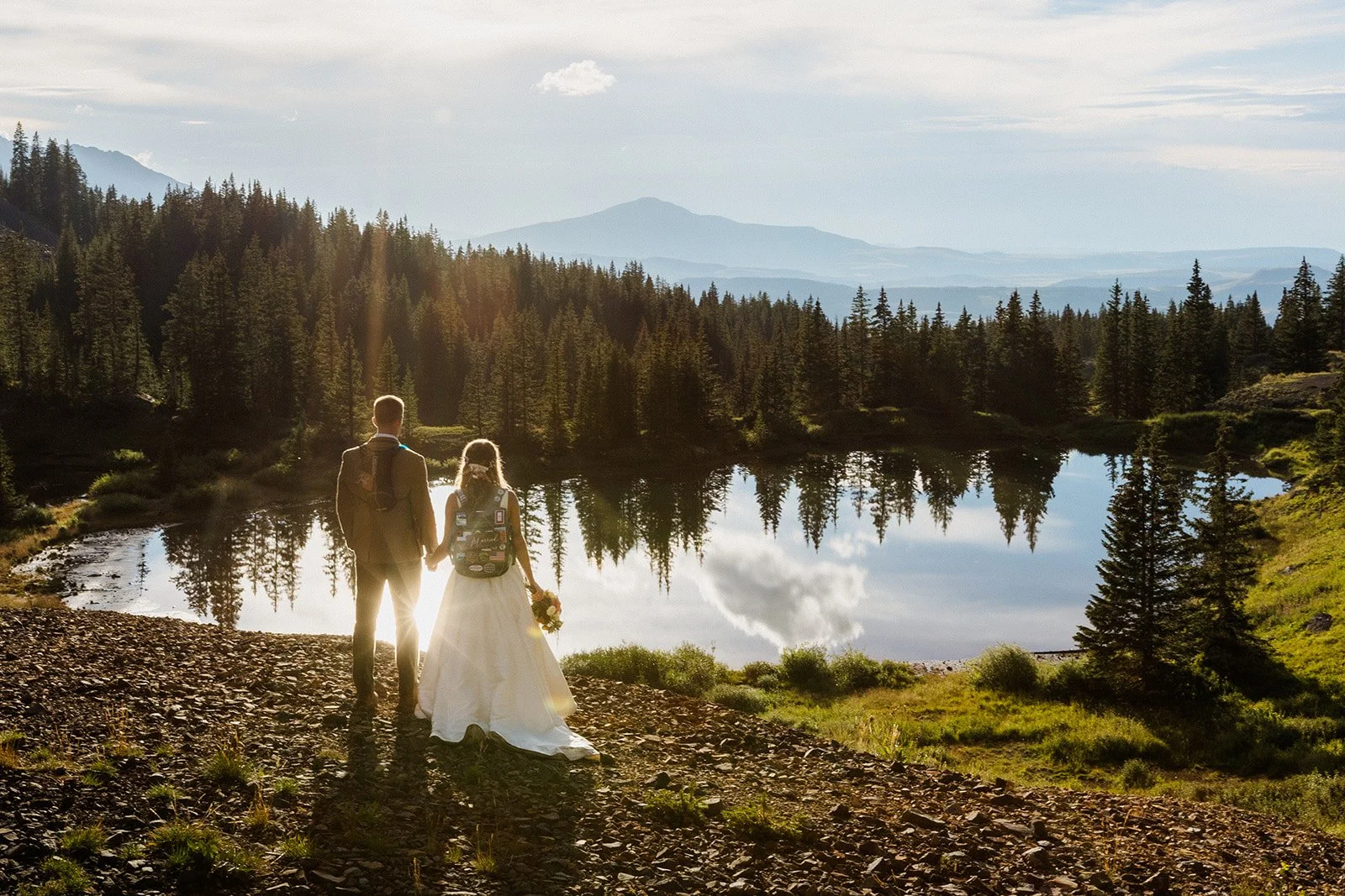 A couple stands in front of a lake outside of Denver in wedding attire with hiking backpacks on holding hands