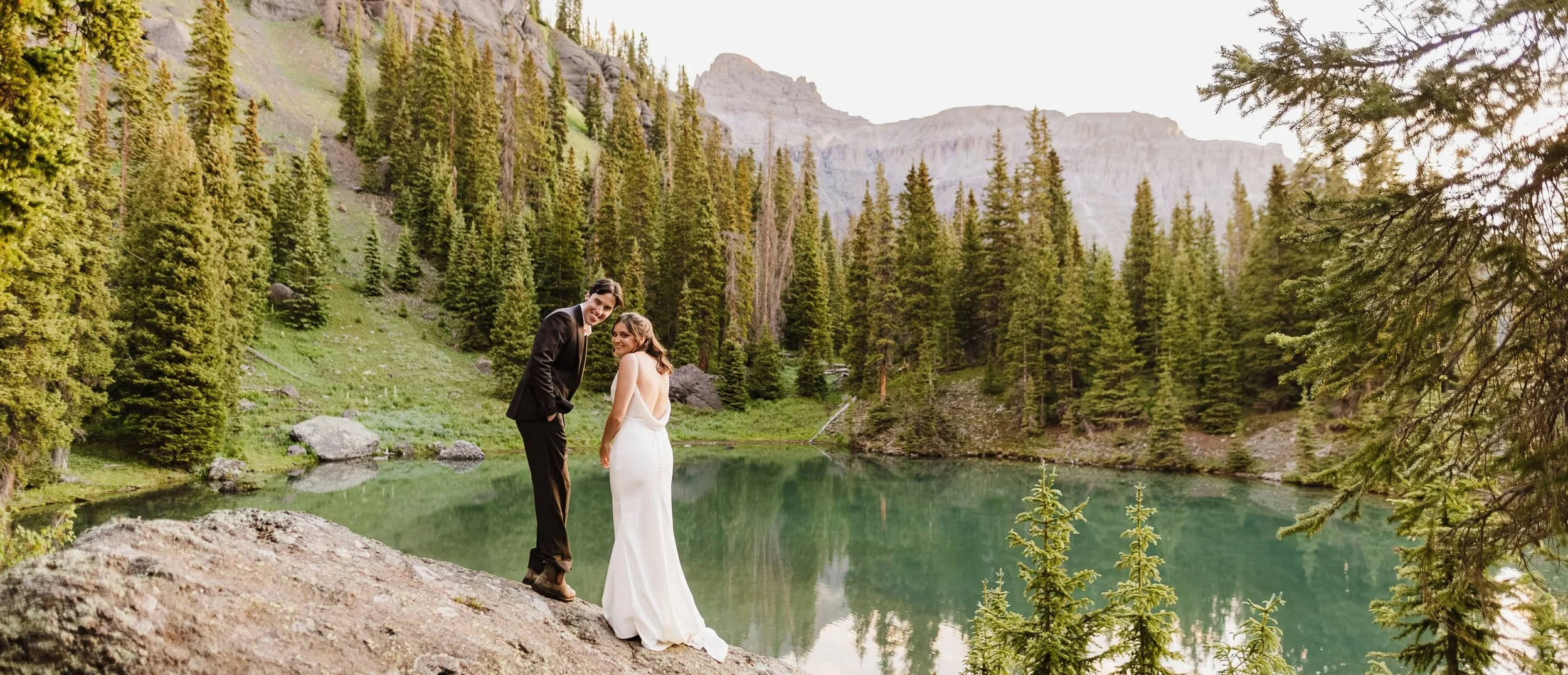 A couple in wedding clothes smiling back at the camera in front of pine trees and a small pond on their Ouray, Colorado elopement day
