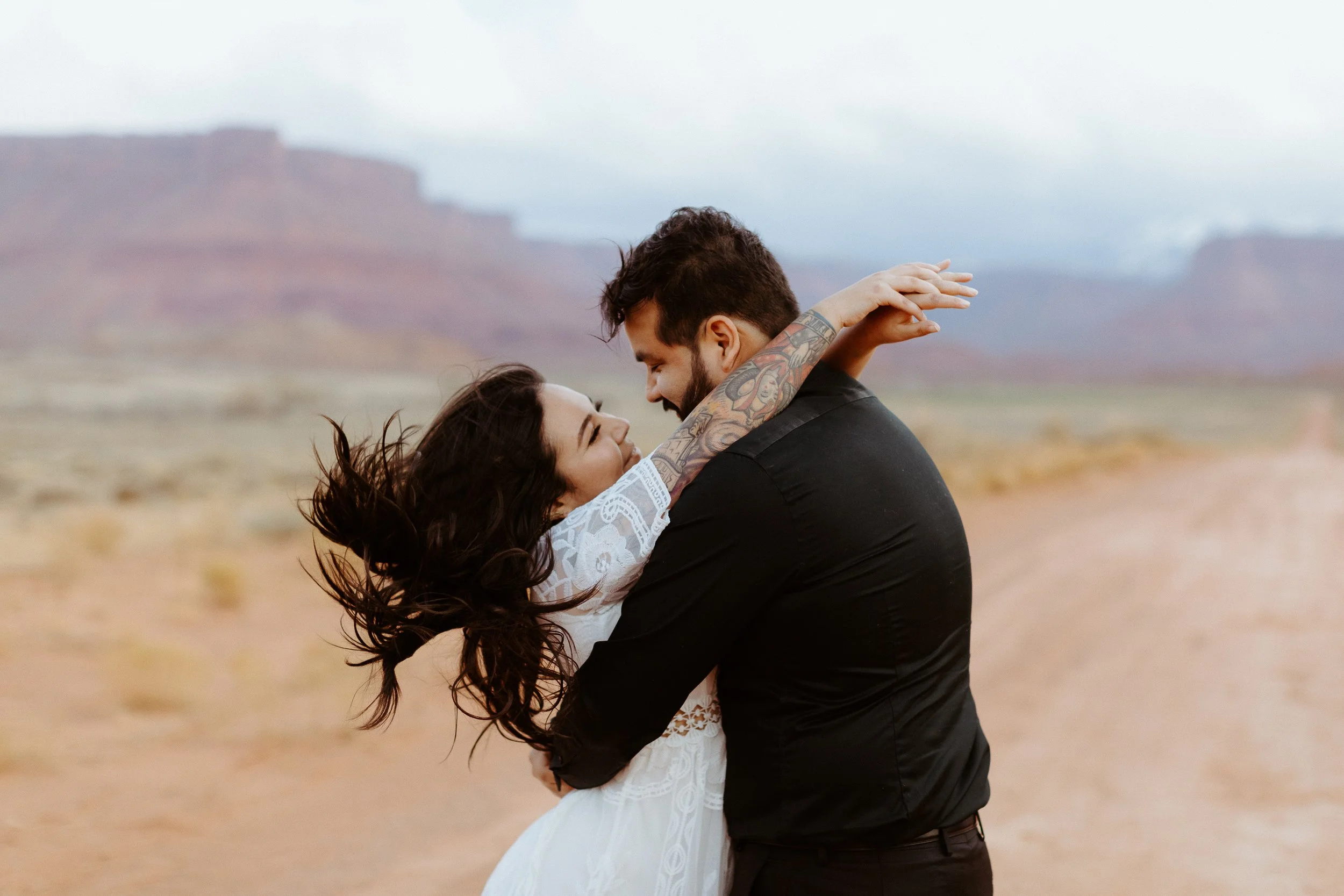 A couple is dancing and embracing in a desert landscape with distant mesas and cloudy sky in the background.
