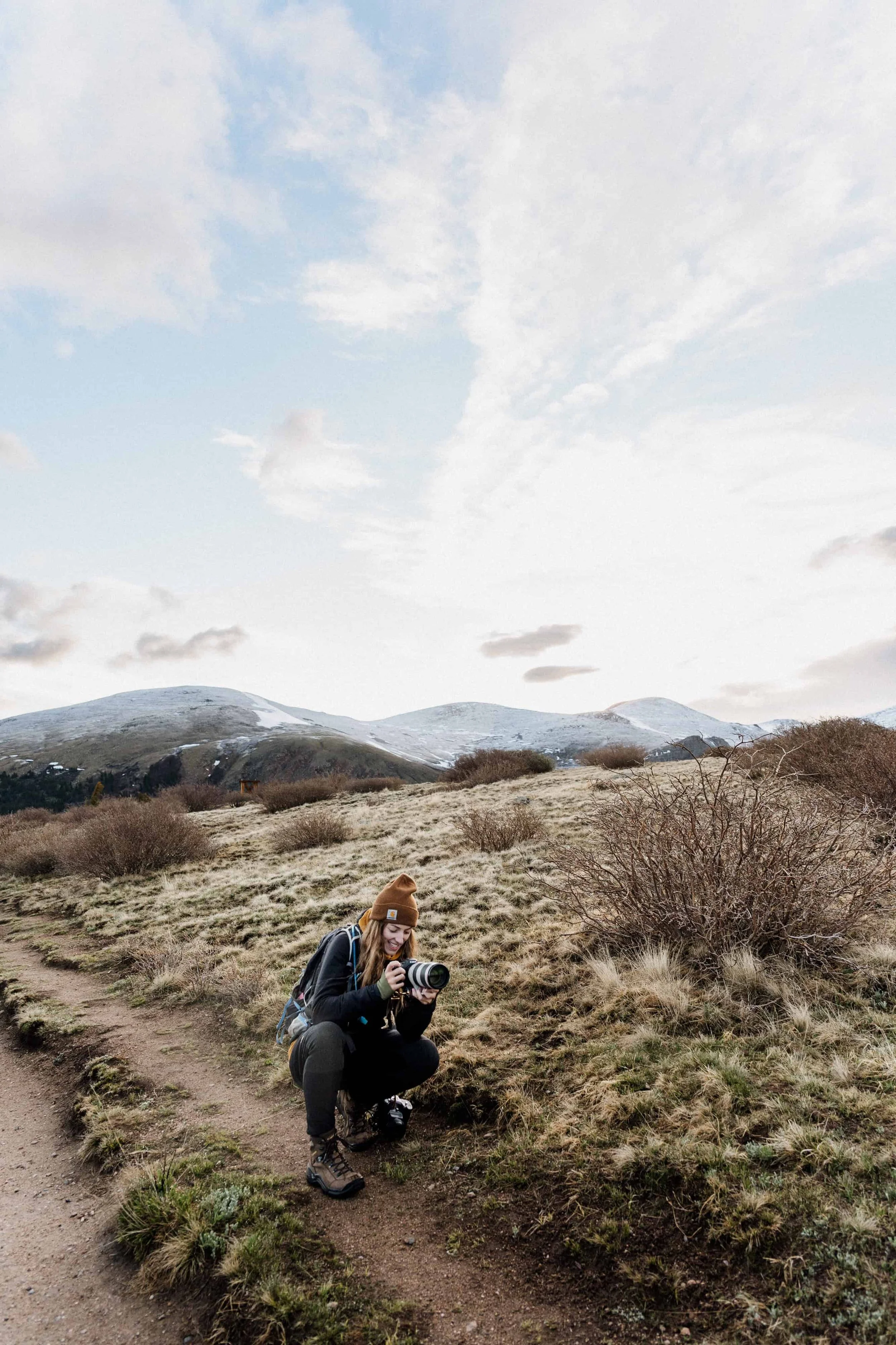 A woman crouching on a trail in a grassy, hilly landscape, holding a camera and smiling, with mountains, cloudy sky, and sparse vegetation in the background.