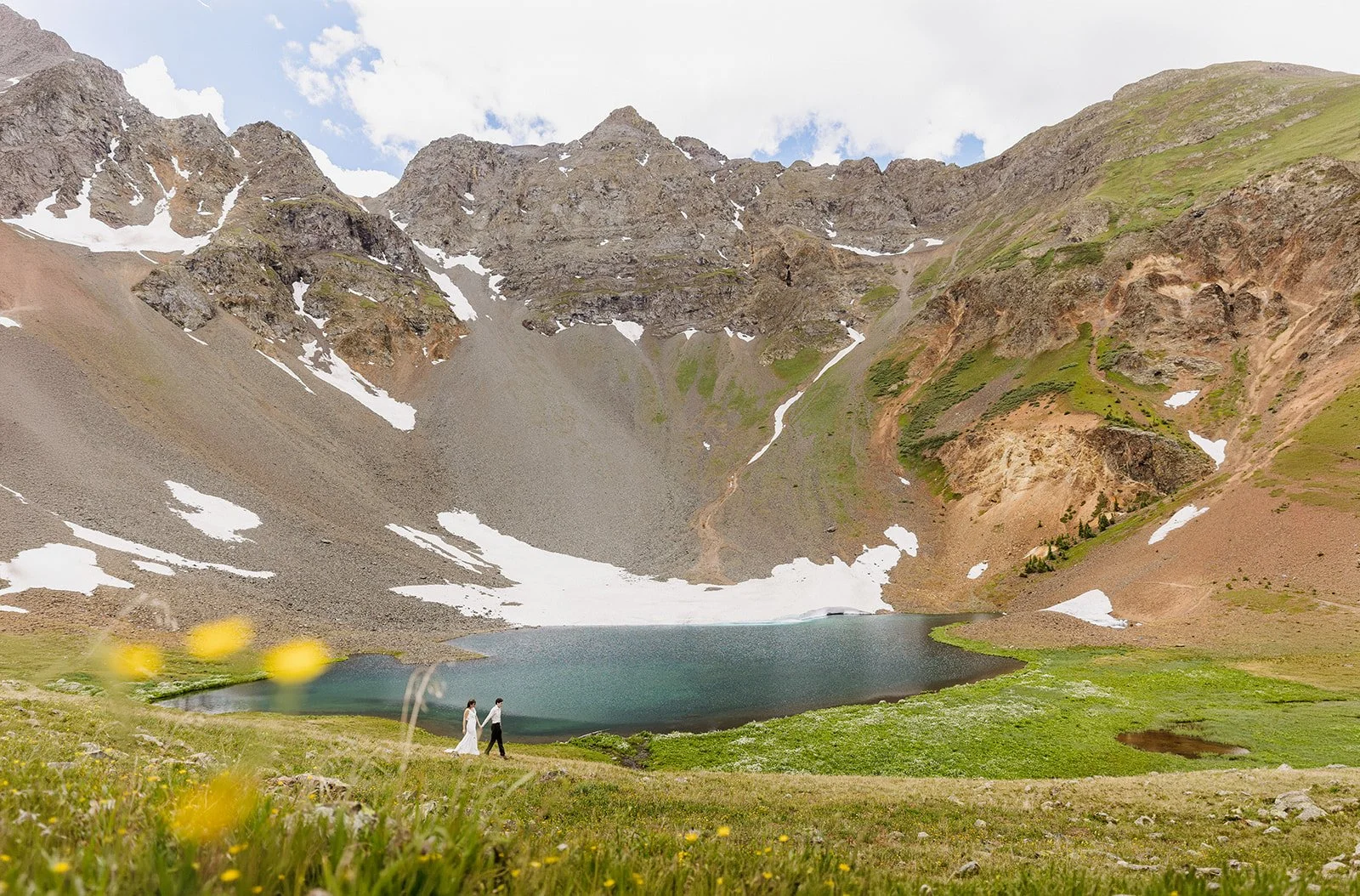 A couple in wedding clothes walks in front of a Turquoise lake outside of Ouray