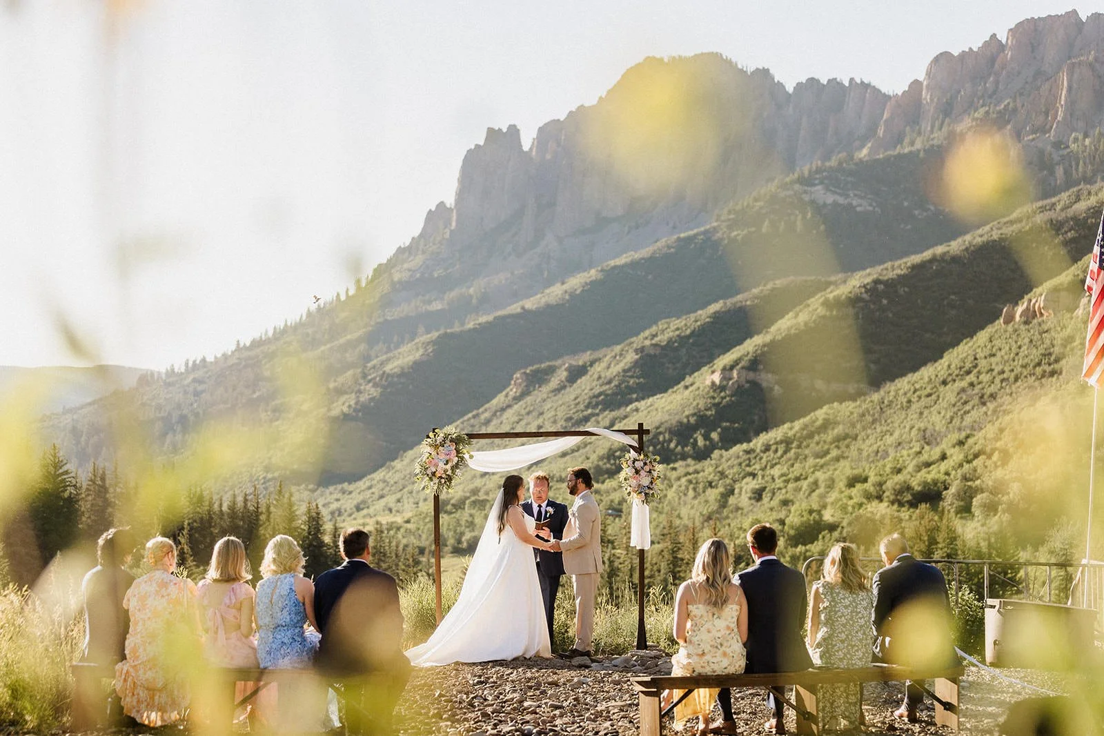 A wedding ceremony taking place outdoors with a mountain backdrop. The bride and groom are standing under a wooden arch decorated with flowers, with an officiant between them. Guests are seated on benches, watching the ceremony. The scene is bathed in warm sunlight, with a flag visible on the right side of the image.