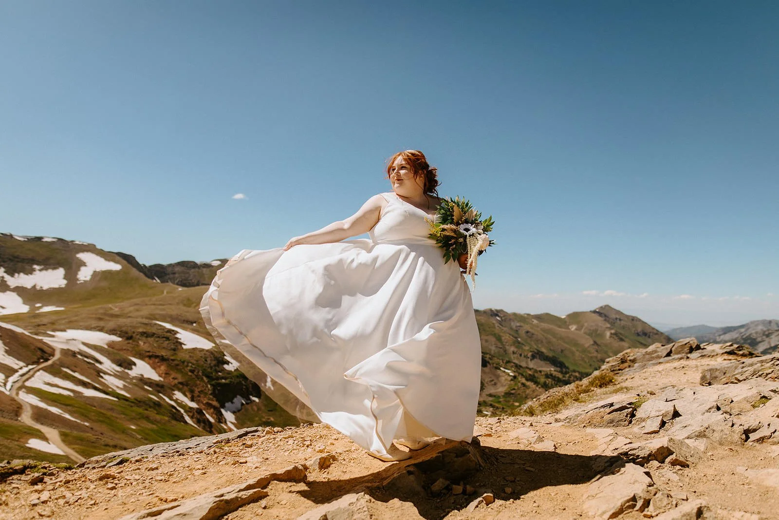 White wedding dress skirt blowing in the wind on a rocky mountain pass with patches of snow