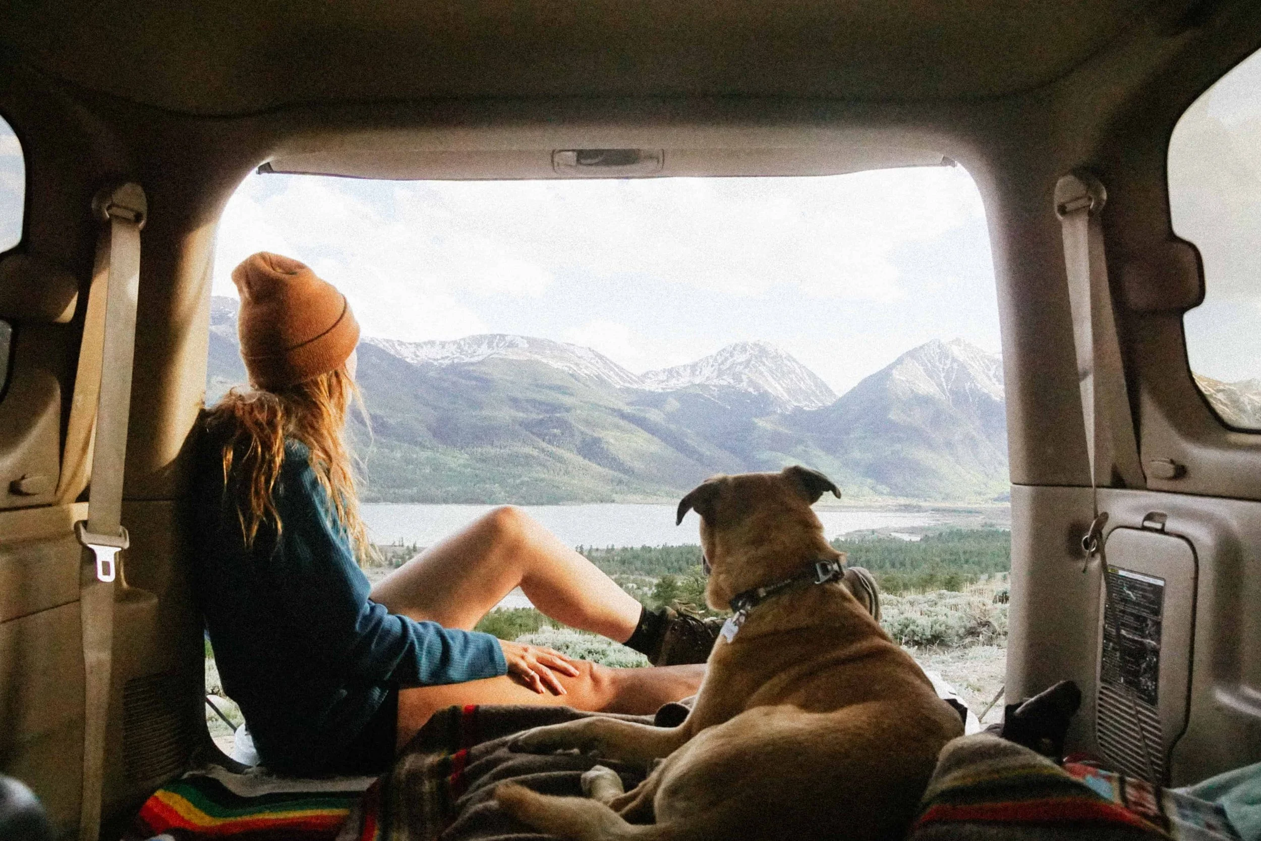 A young woman with long hair, wearing a beanie, sitting inside a vehicle with her dog, enjoying a mountain landscape with snow-capped peaks and a lake visible through the open rear of the vehicle.