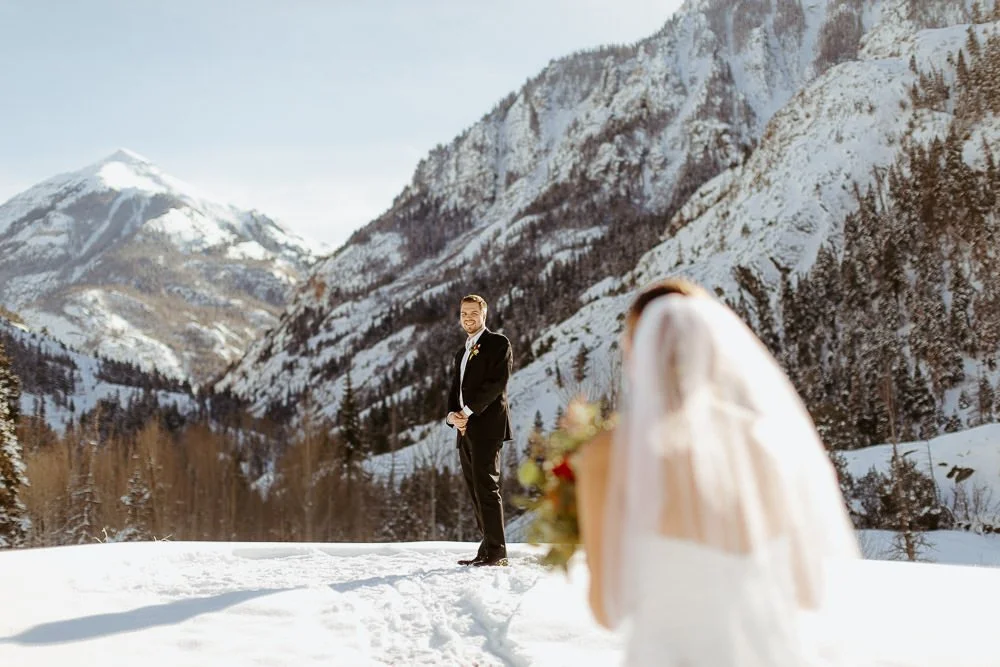 A bride in a long veil looks back at her groom in front of snowy cliffs in Ouray